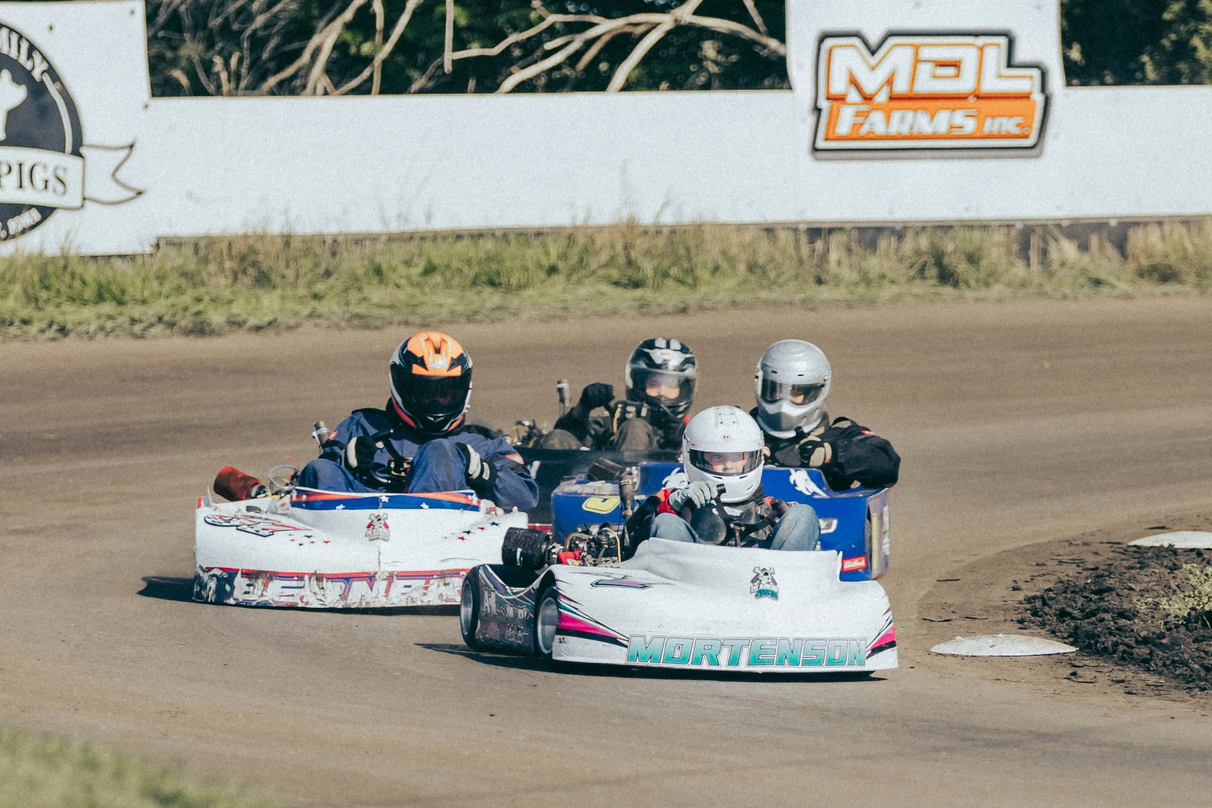Four go-kart racers on a dirt track, all wearing helmets and racing suits, with a billboard and grass in the background.