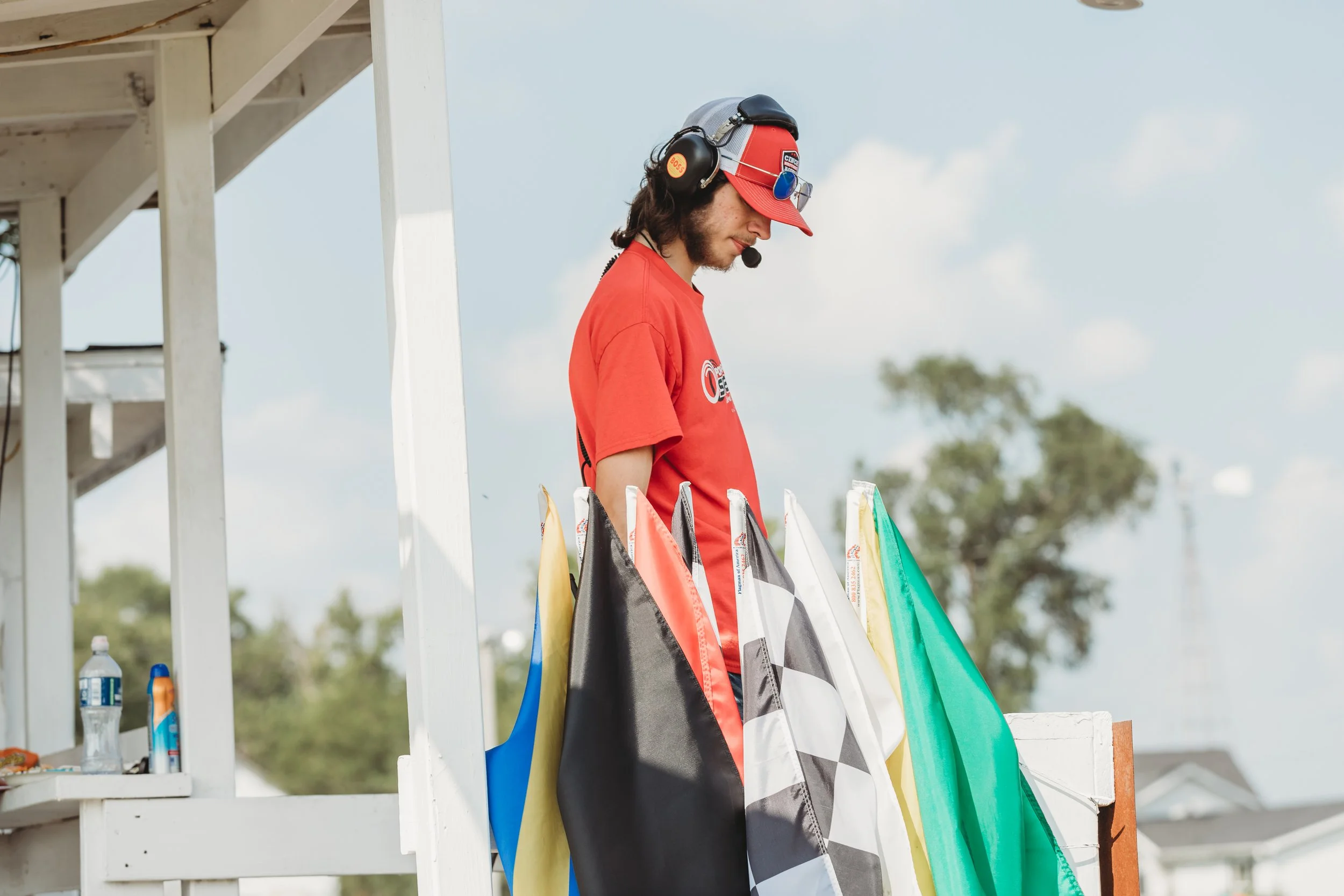 A man wearing a red T-shirt, a red and gray cap, and a headset with a microphone, standing at a white booth with colorful flags hanging on the side, outdoors on a sunny day with trees and buildings in the background.