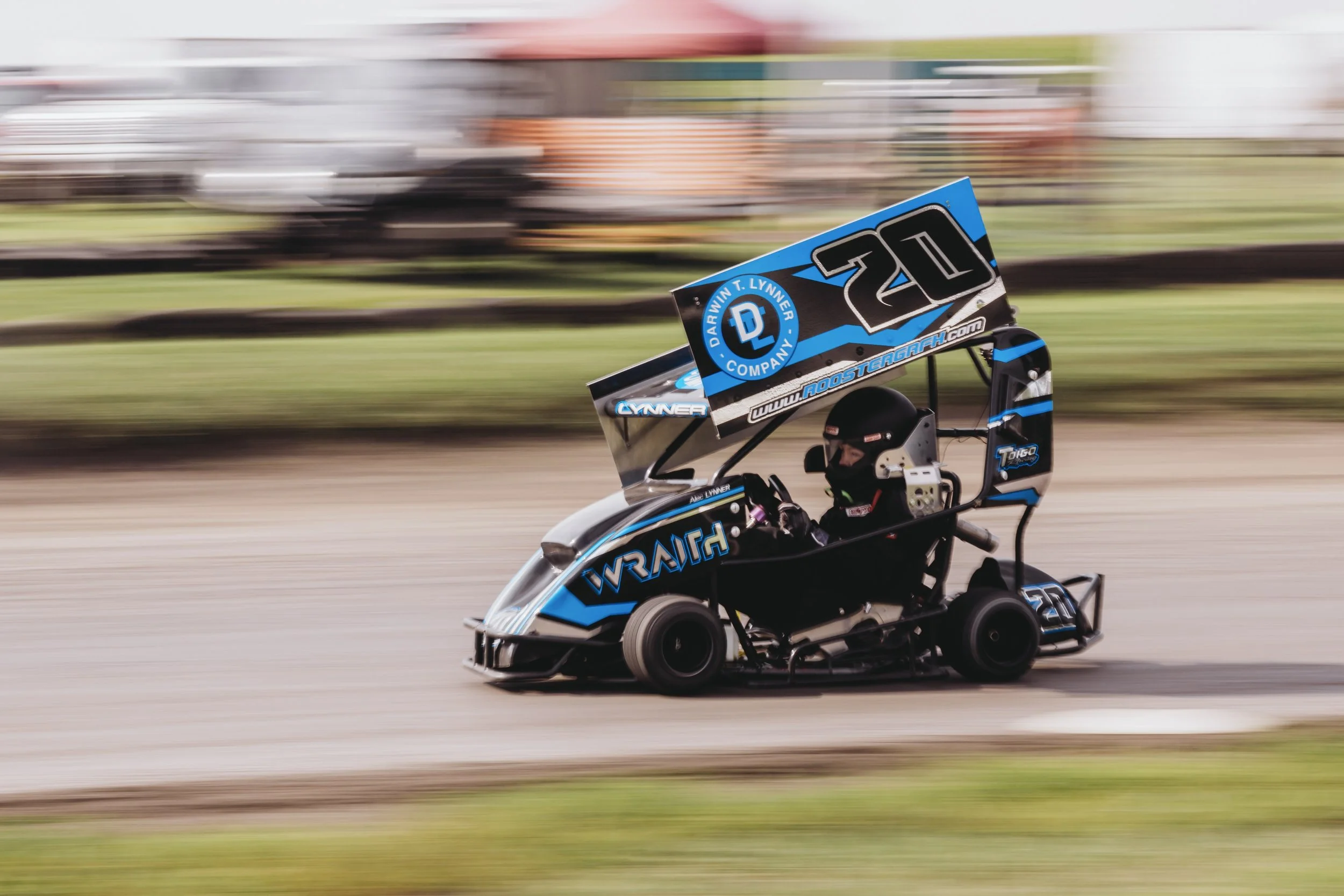 A small race car with blue and black graphics speeds on a dirt track, driven by a person in a black helmet and racing suit.