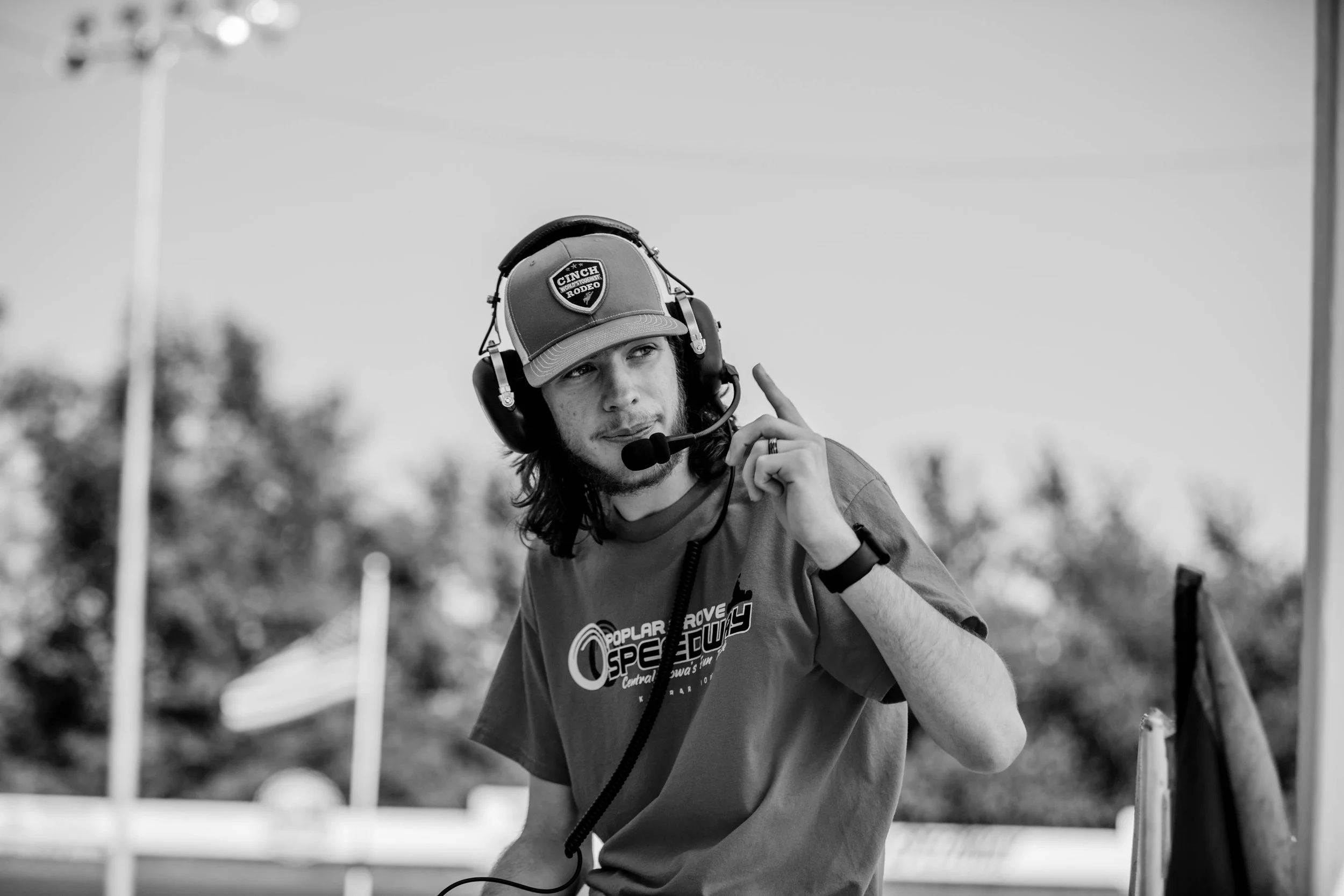 A man wearing a headset with a microphone and a baseball cap, pointing upwards while standing outdoors.