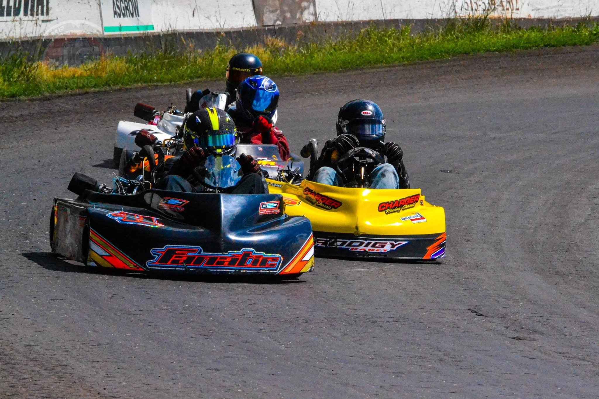 Two racing go-karts with drivers wearing helmets and racing suits on a dirt track, closely competing.