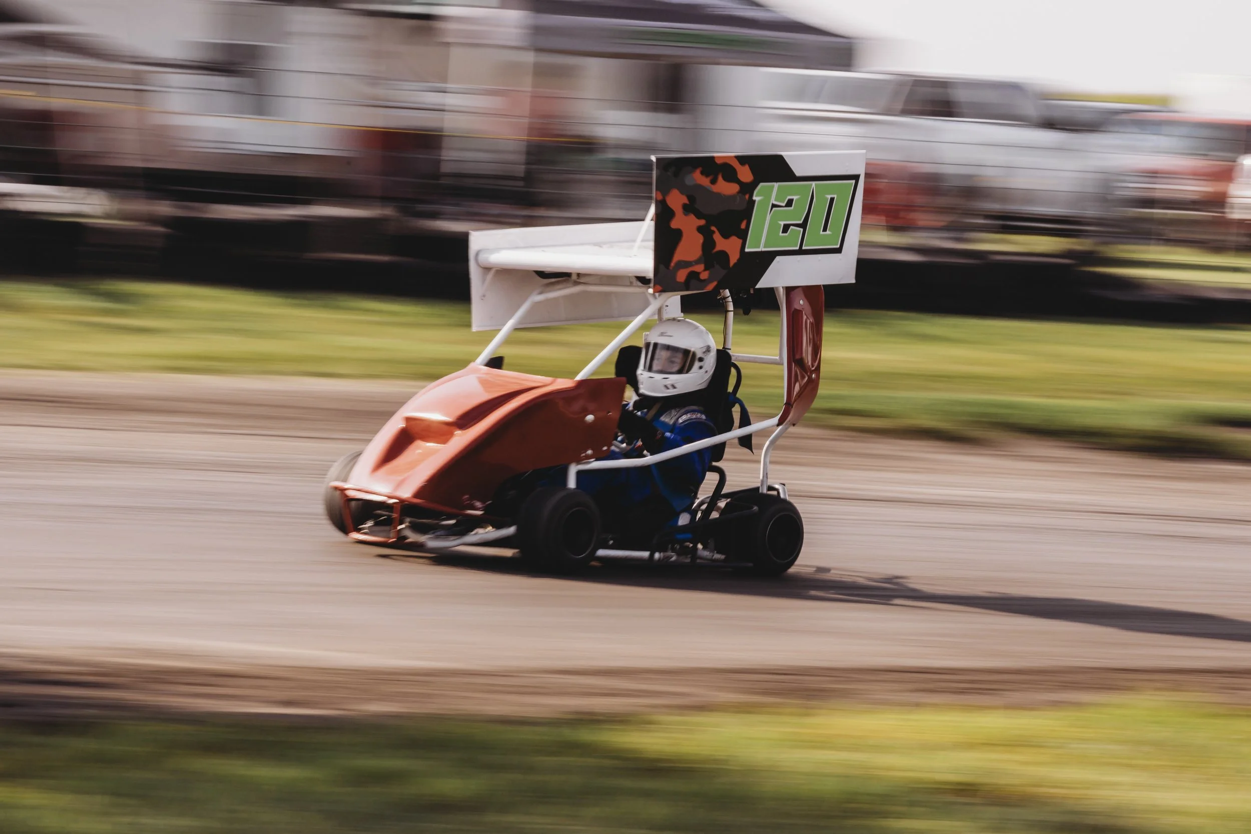 A small race car, number 120, is speeding on a dirt track with motion blur in the background. The driver, wearing a white helmet and blue racing suit, is seated inside.