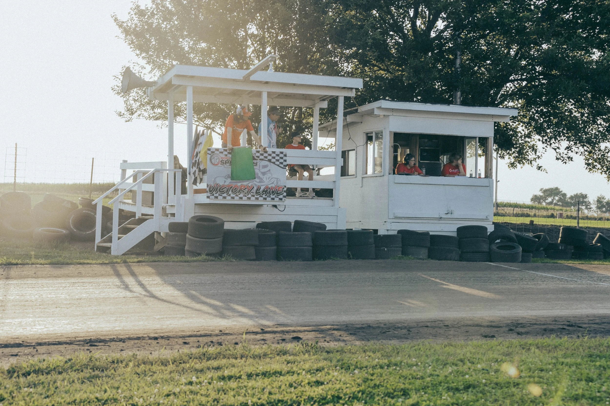 Race control tower and spectators at a Speedway dirt track race, with tires lining the track and a sign that reads 'Victory Lane'.