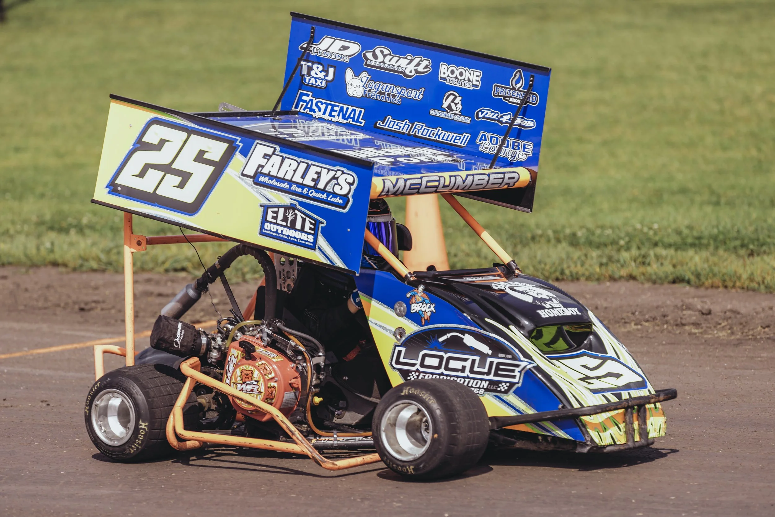 A small racing go-kart on a dirt track with a colorful blue, yellow, and black body, large rear wing with sponsor logos, and racing tires.