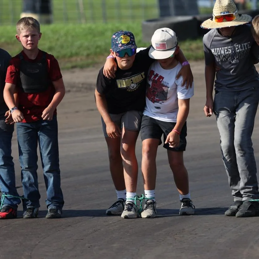 Group of children standing side by side on a race track, with two kids in the middle helping each other to walk.