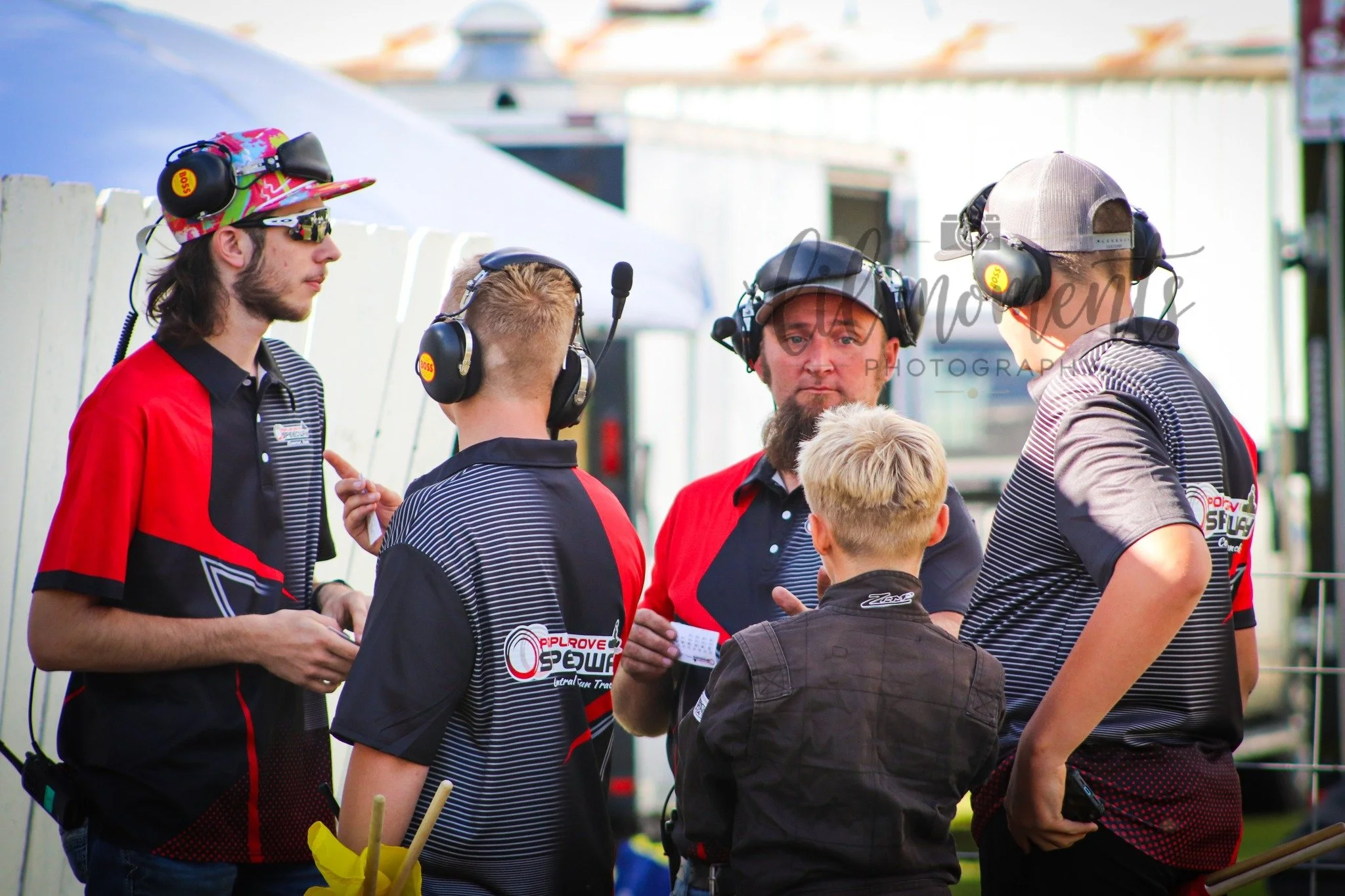 Group of people, including technicians and team members, wearing headsets and team uniforms, engaged in a discussion at a racing event.