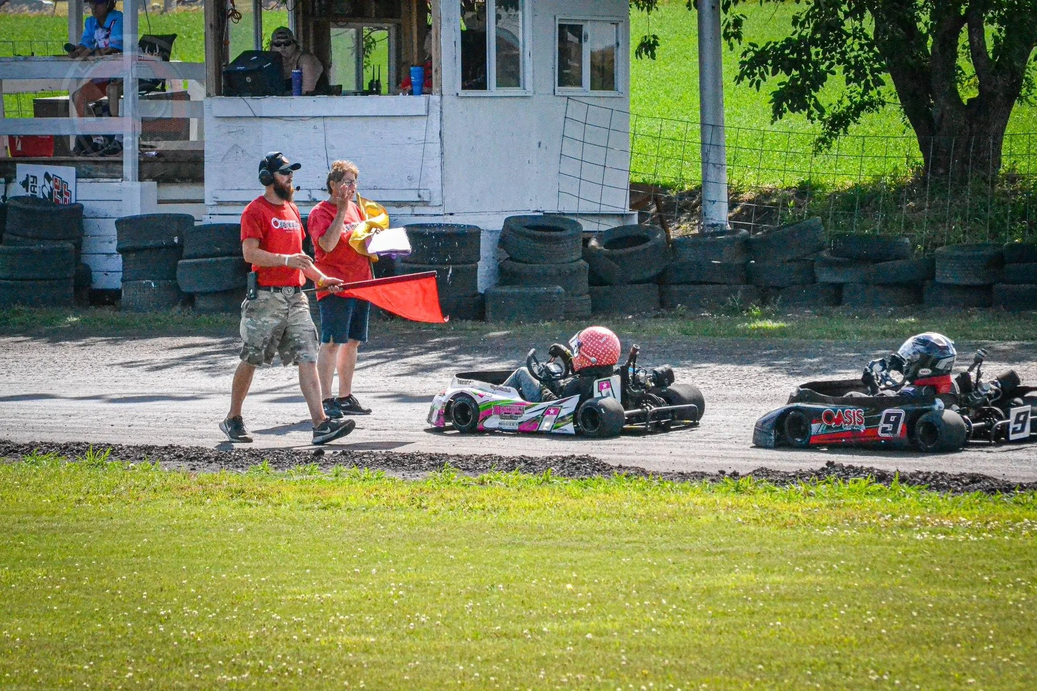 Two go-karts on a race track, with two officials standing nearby, one holding a red flag. In the background, there is a building with people and equipment inside, and tires stacked against a fence. The setting appears to be an outdoor go-kart racing event.