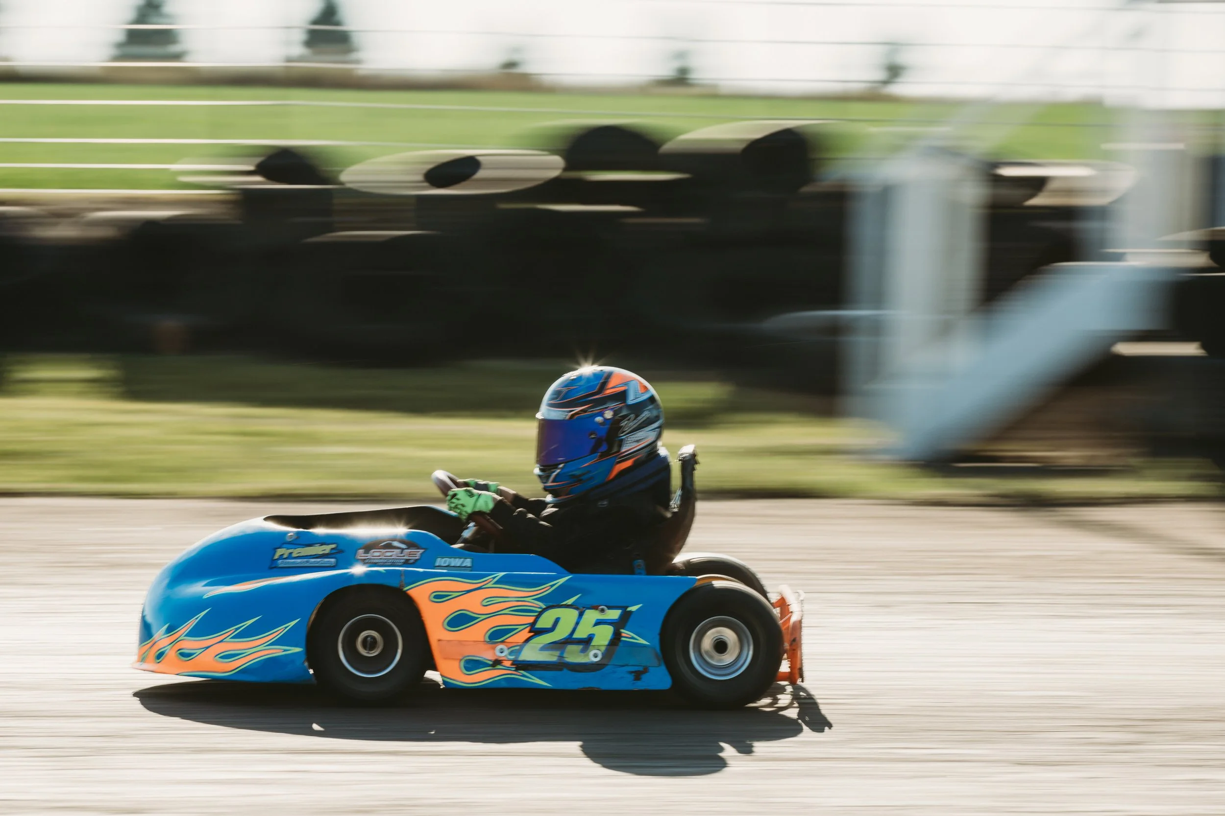 A young person driving a go-kart on a racing track, wearing a blue helmet and black racing suit, with a blue and orange go-kart numbered 25.