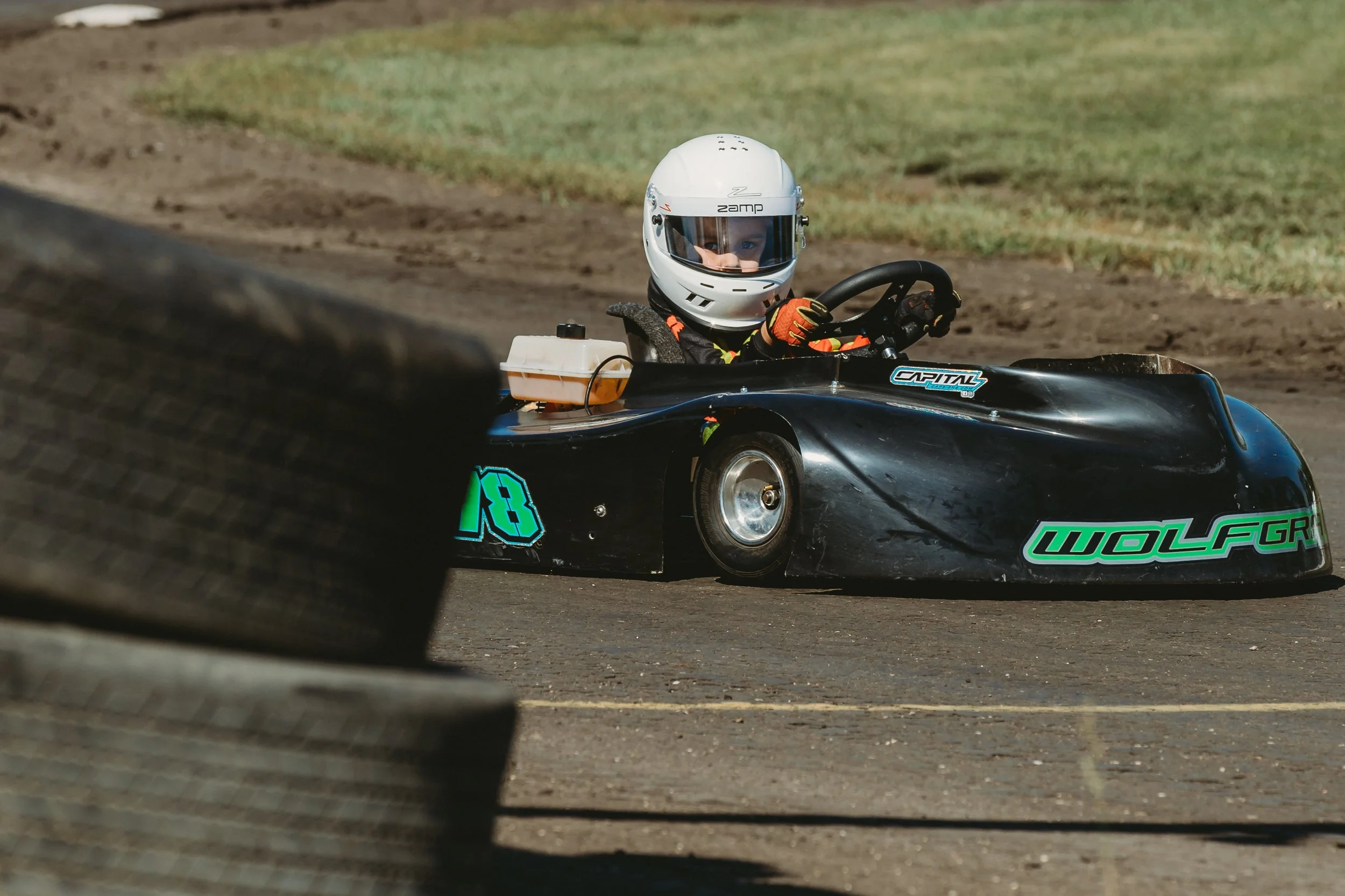 Young boy driving a black go-kart with the number 18, wearing a white helmet and racing gloves on a dirt track.