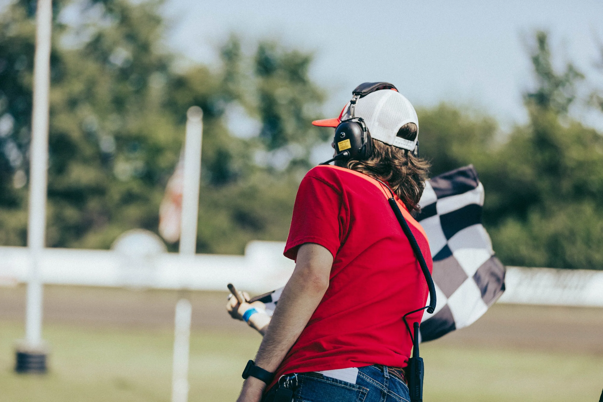 A person with long hair wearing a red shirt, gray cap, and headphones holding a radio device and a checkered flag, outdoors on a grassy field with trees in the background.