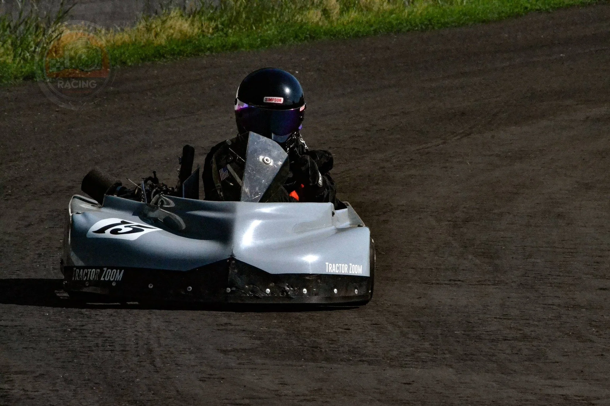 A person wearing a black helmet and racing suit operates a small, silver racing vehicle with the words 'Tractor Zoom' on the side, on a dirt track at night.