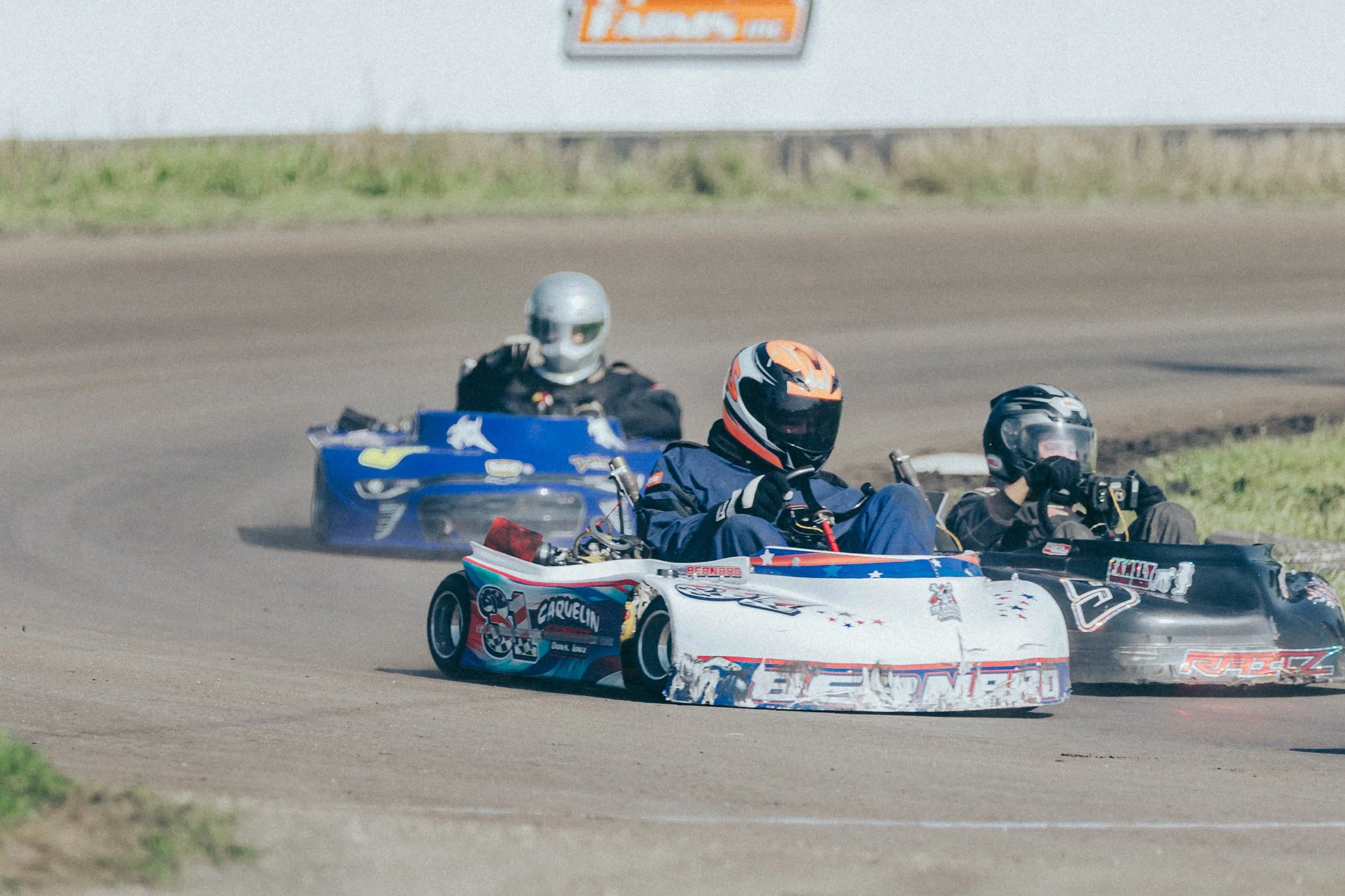 Three mini cars race on a dirt track, each driven by a person wearing a helmet and racing gear.