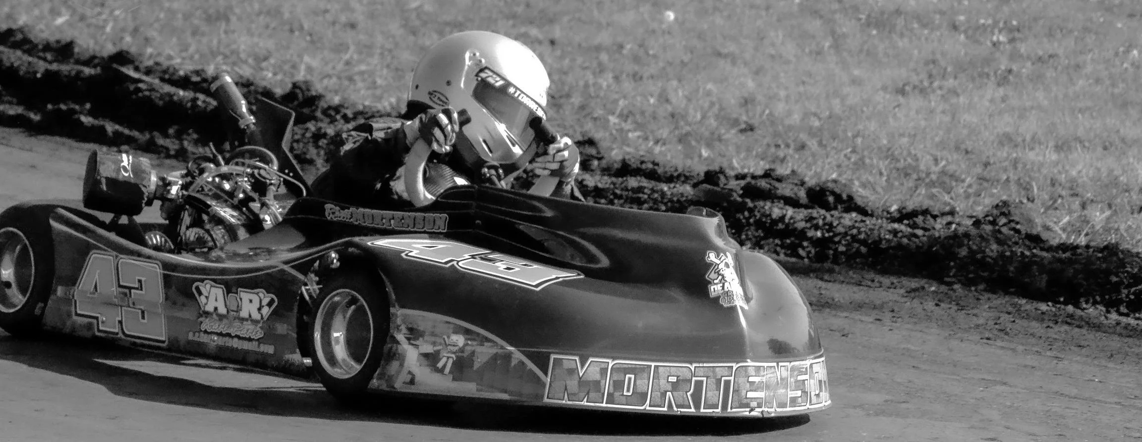 A race car driver in a helmet, sitting in a black race car on a dirt track.