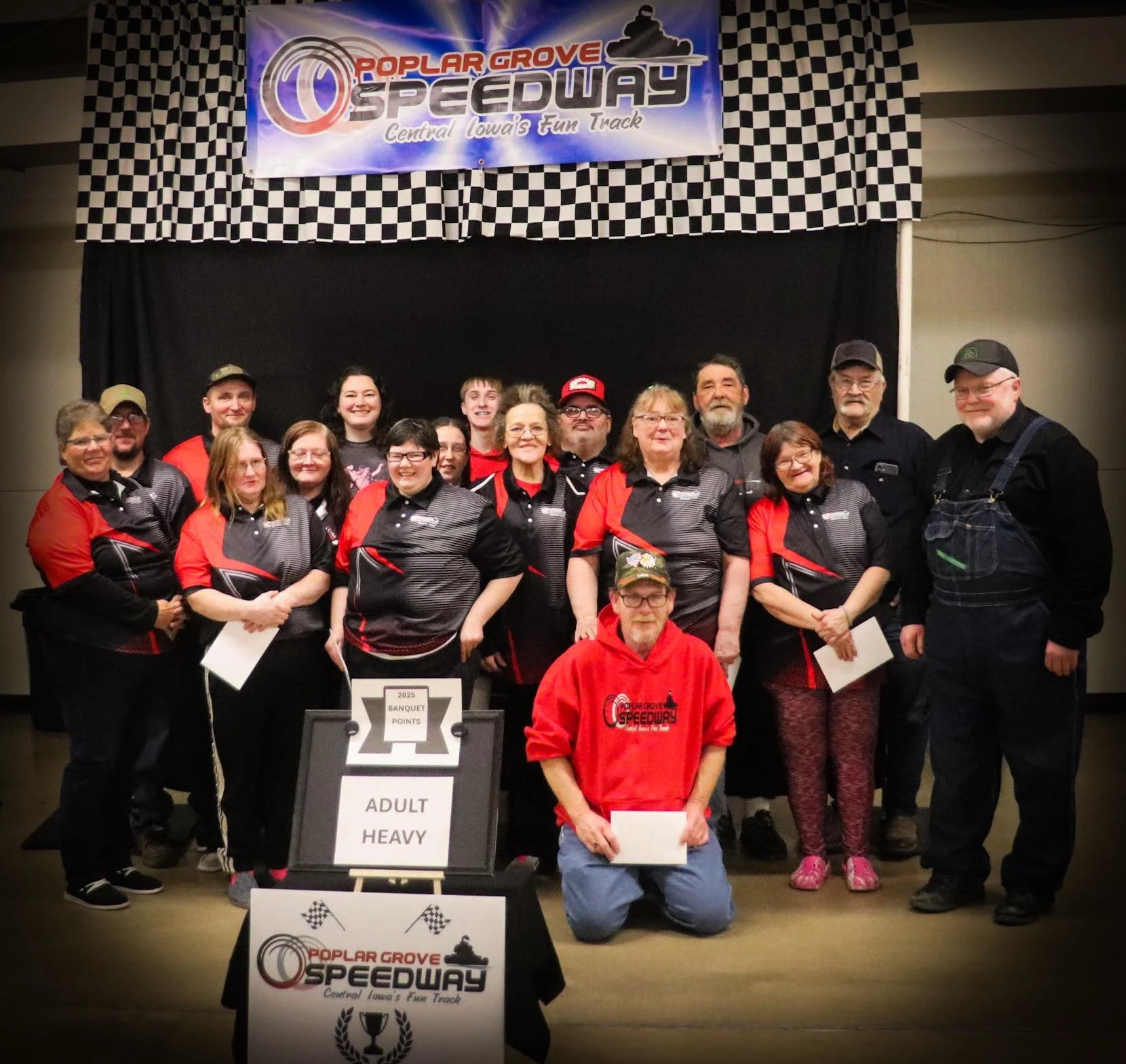 Group of people at a speedway event in front of a banner that reads 'Poplar Grove Speedway Central Iowa's Fun Track,' with a table displaying a sign that says 'Adult Heavy' and a trophy illustration.