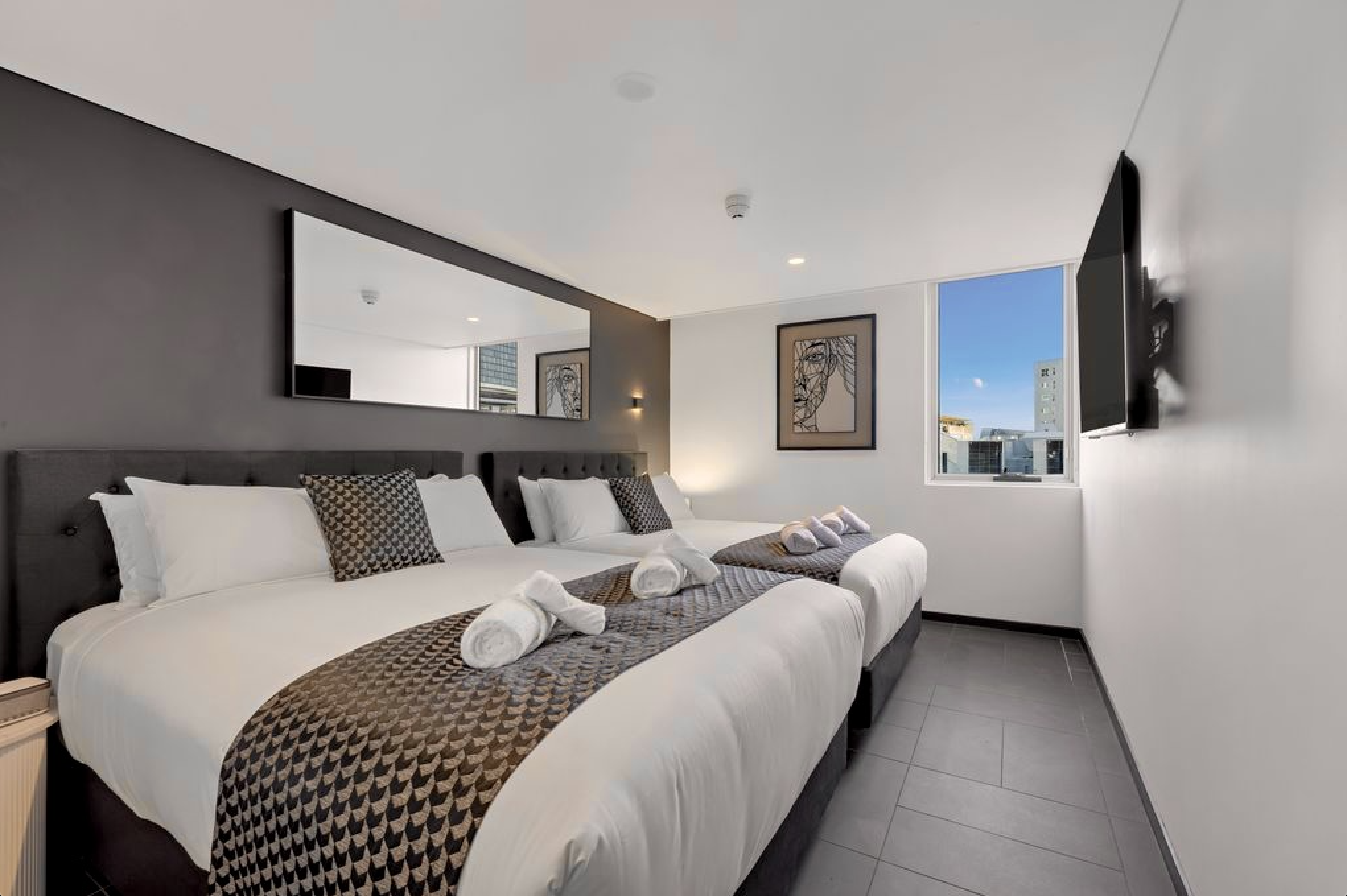Modern hotel room with two beds, black upholstered headboards, decorative pillows, a large mirror, artwork on the wall, a window with city view, and a wall-mounted flat screen TV.