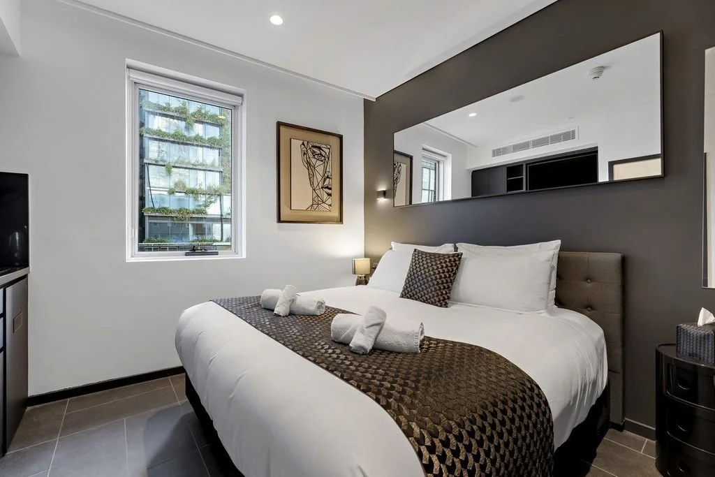 Modern hotel bedroom with a large bed, white bedding, and a patterned brown bed runner. The bed has four rolled towels and a decorative pillow. Two windows let in natural light. A large mirror spans the width of the dark accent wall behind the bed, with a framed minimalist line art piece on the wall beside it. There is a small nightstand with a lamp on each side of the bed.