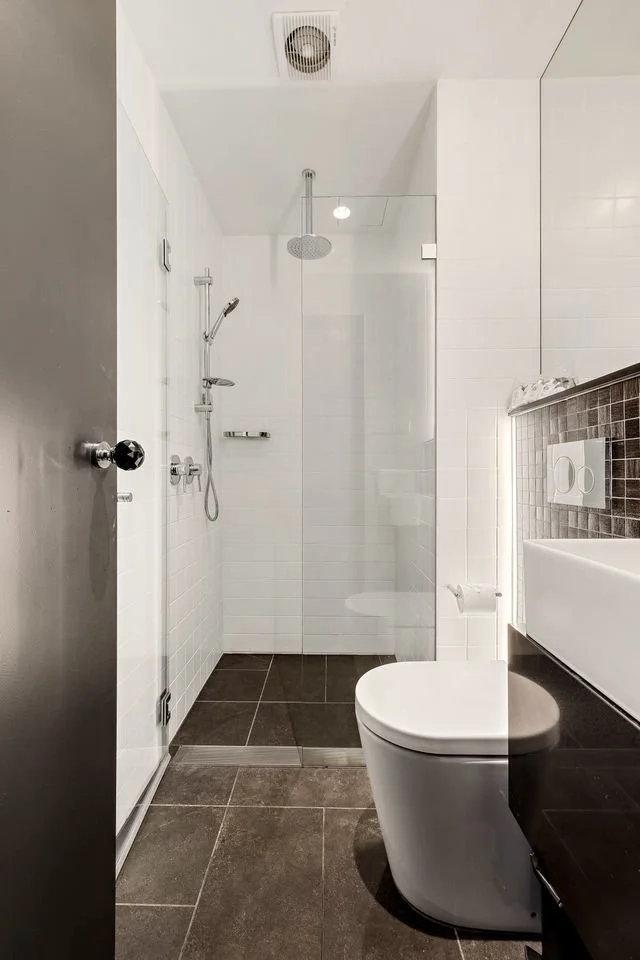 Modern bathroom with a walk-in shower enclosed by glass, a white toilet, and a white wall-mounted sink. Dark floor tiles and white wall tiles, with a mirror above the sink and ceiling ventilation.