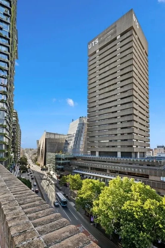 Cityscape featuring a tall concrete building with 'UTS' on top, other modern buildings, a street with cars and a bus, and green trees along the sidewalk under a clear blue sky.