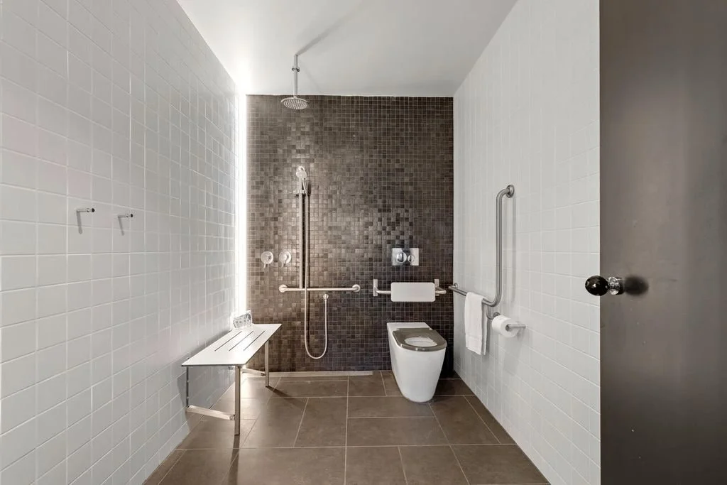 Accessible bathroom with brown tiled floor, white and dark brown tiled walls, a raised toilet with handrails, a shower area with a handheld showerhead, towel holder, and a bench.