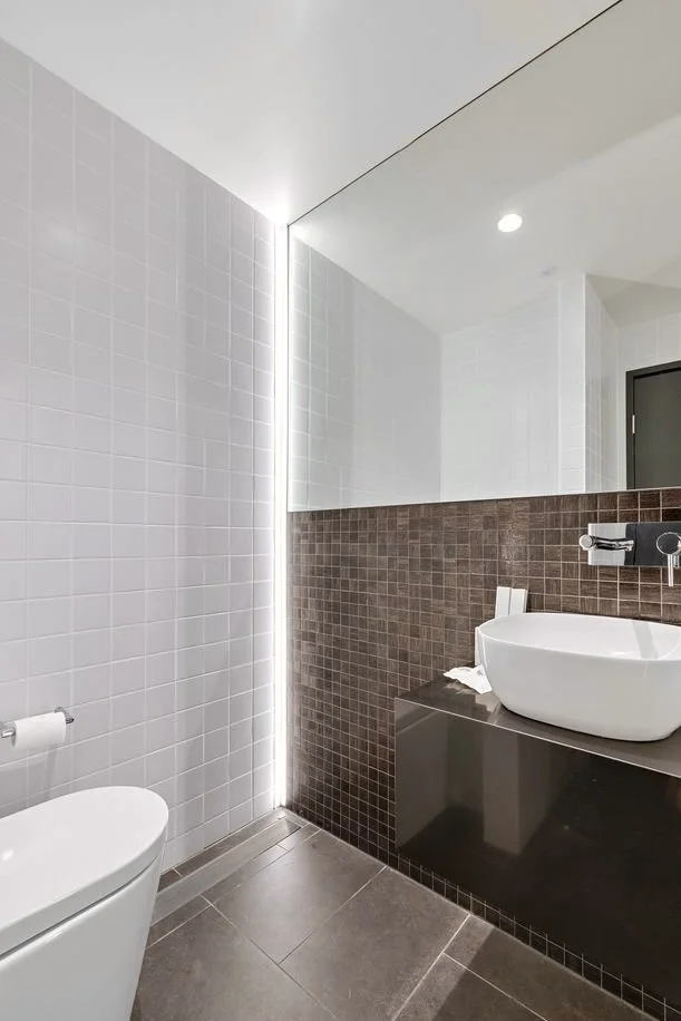 Modern bathroom with white tiled wall, brown tiled half-wall, large mirror, white vessel sink on black vanity, and toilet with toilet paper.