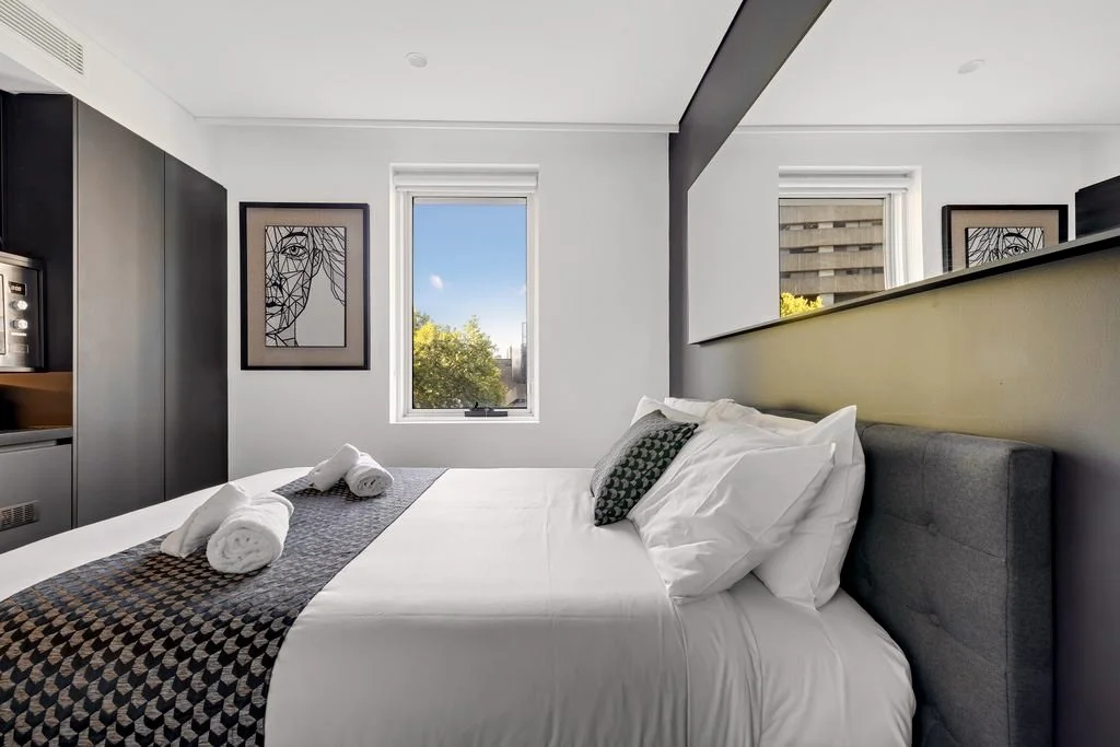 A modern bedroom with white walls, a large bed with white linens and a patterned pillow, two rolled towels on a runner, and artwork of an abstract face on the wall. There are two windows showing trees and a building outside.