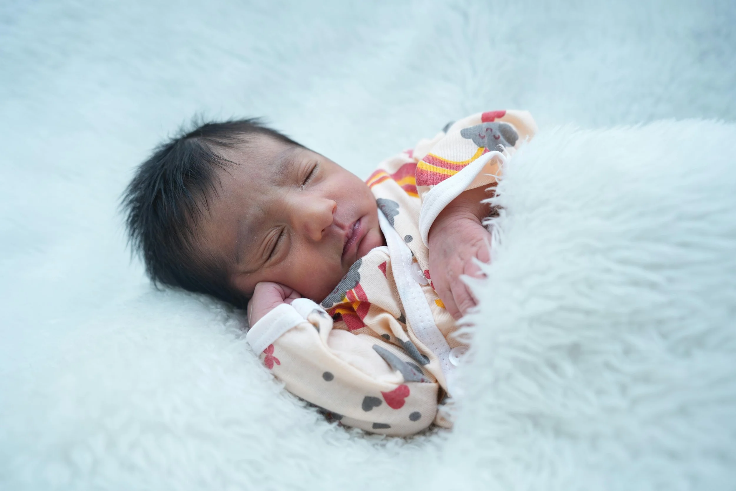 A sleeping baby lying on a soft, light-colored blanket, wearing a cream-colored outfit with colorful heart and animal prints.