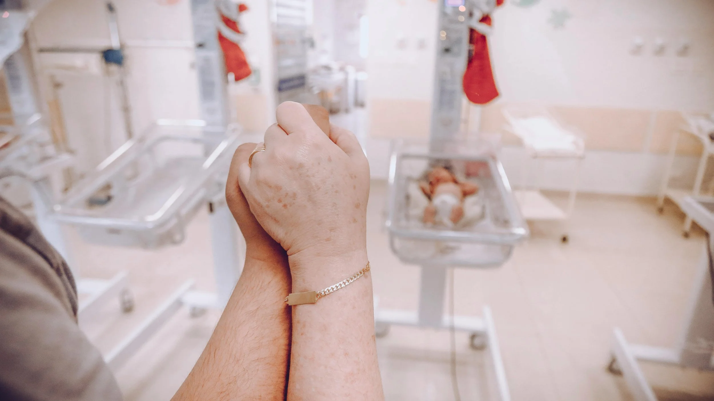 Person holding their hand, with a hospital baby incubator in the background where a baby is lying on a hospital bed.
