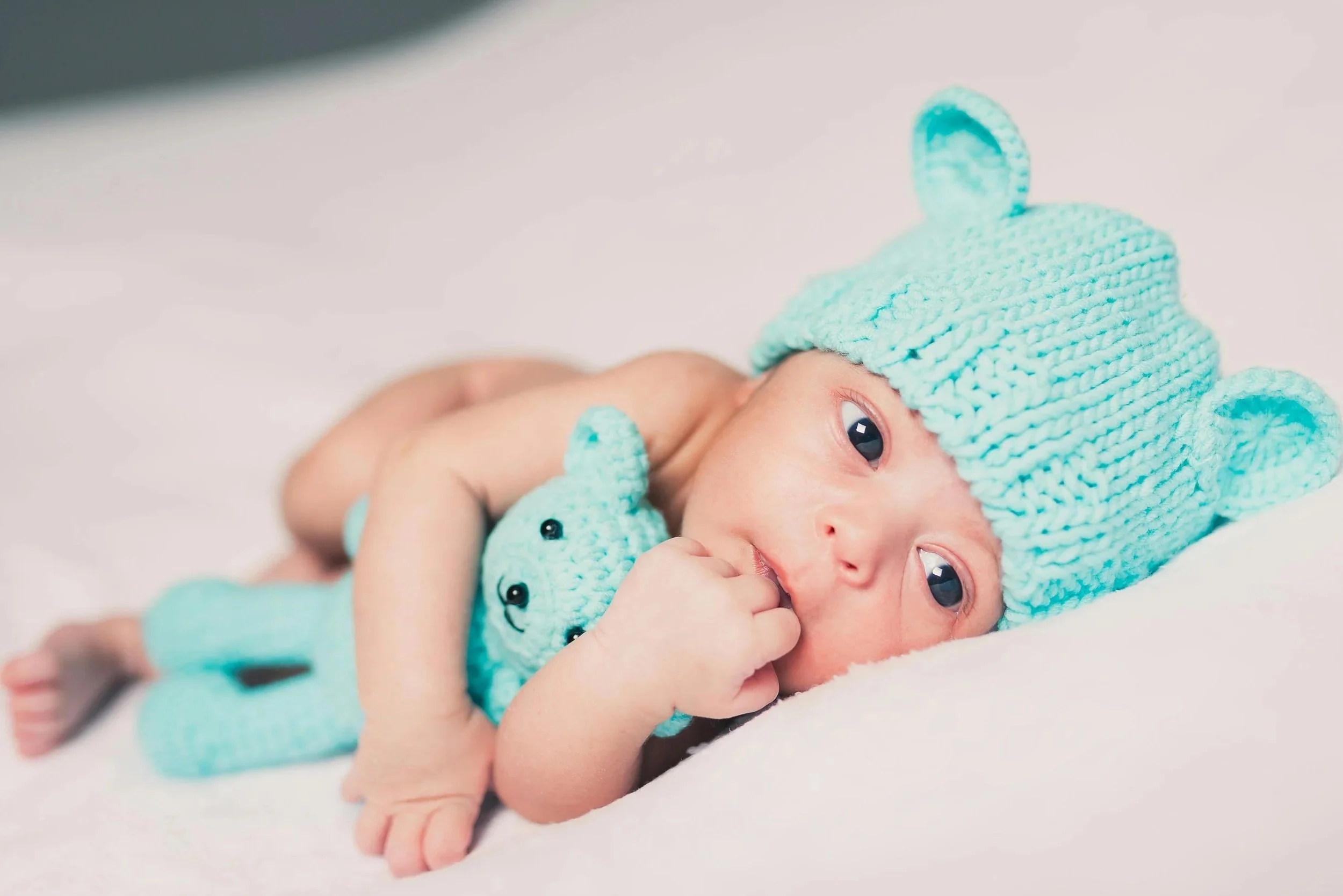 A newborn baby lying on a white surface, wearing a light blue knitted hat with bear ears, holding a small blue teddy bear close to their face.