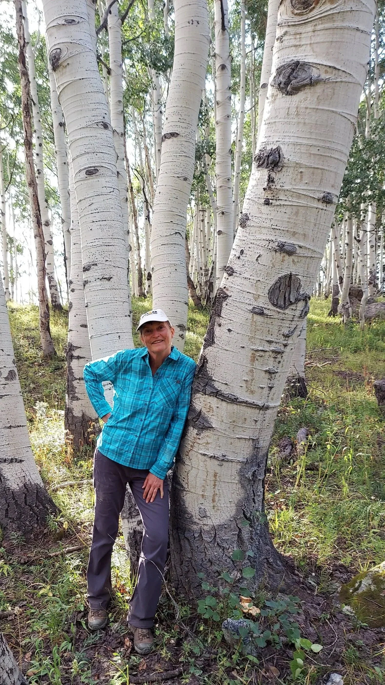 A smiling woman in outdoor hiking attire leaning against a group of tall white birch trees in a forest.