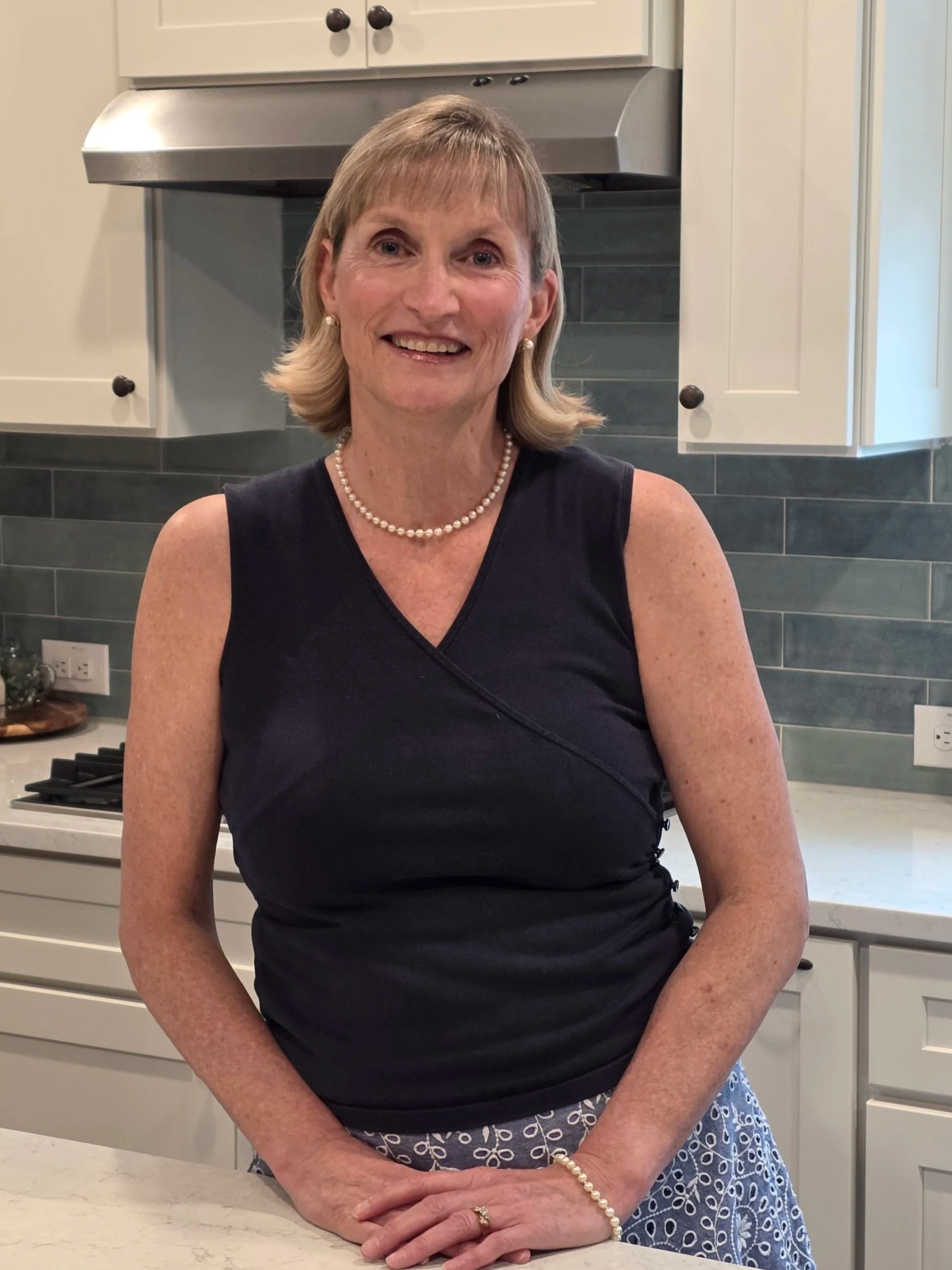 A woman with short, blonde hair smiling at the camera, wearing a black sleeveless top, pearl necklace, and earrings, standing in a modern kitchen with white cabinets, a gray backsplash, and a stove in the background.