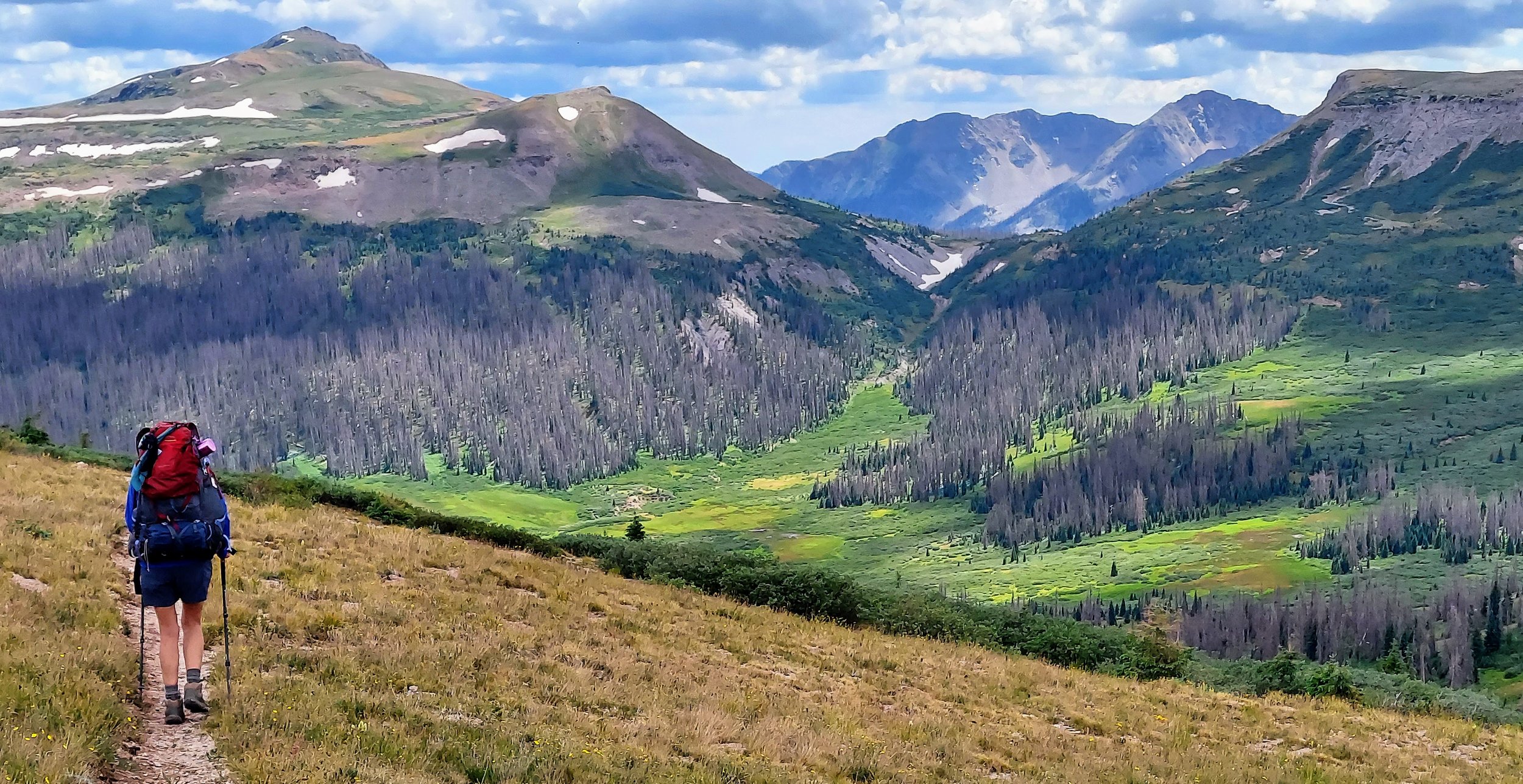 Hiker walking along a trail in a mountainous landscape with green valleys, sparse trees, and snow patches on the mountains under a partly cloudy sky.