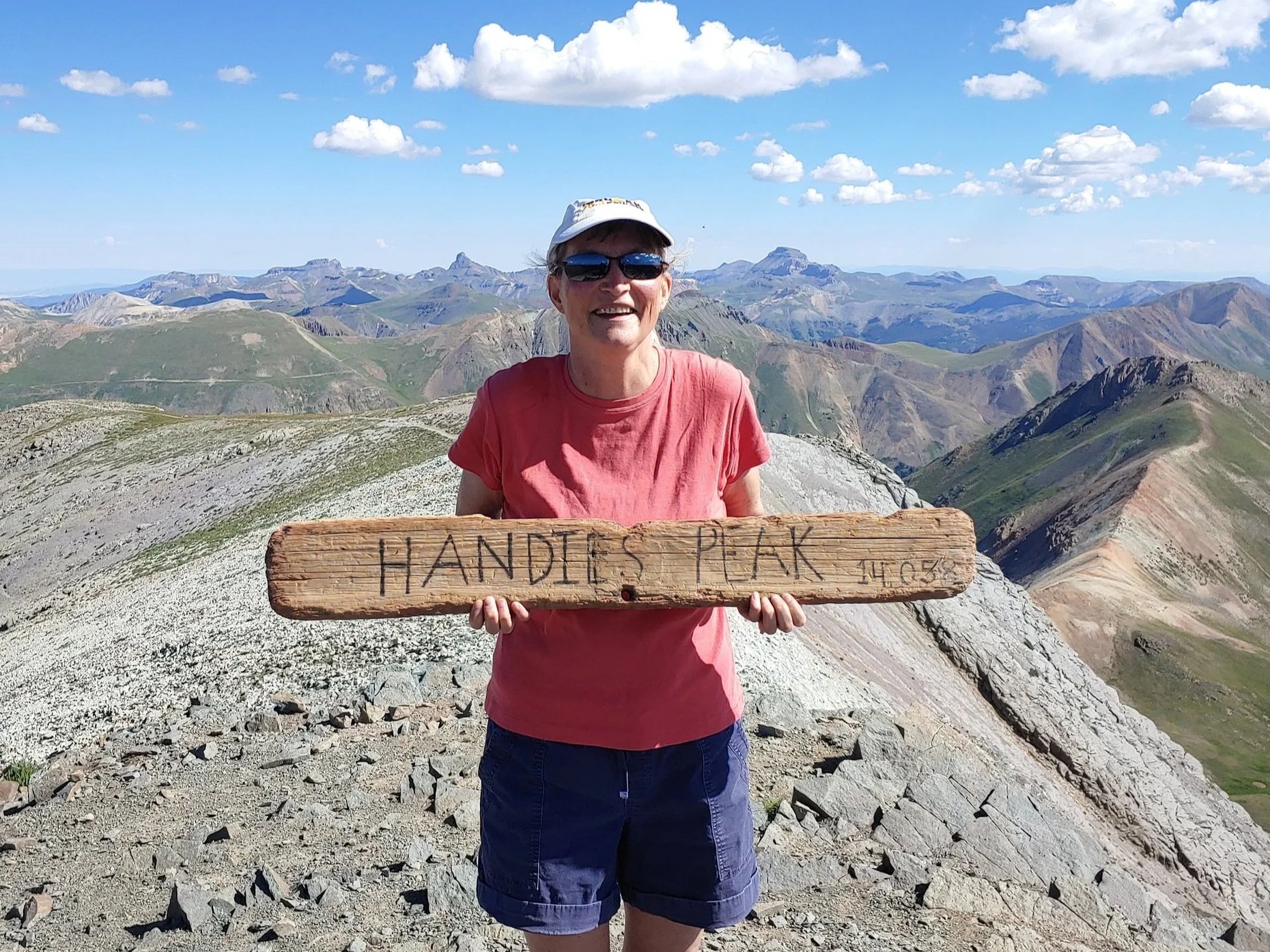 Woman wearing sunglasses and a white hat holding a wooden sign that says "HANDIES PEAK" on a mountain trail with a mountainous landscape and blue sky in the background.