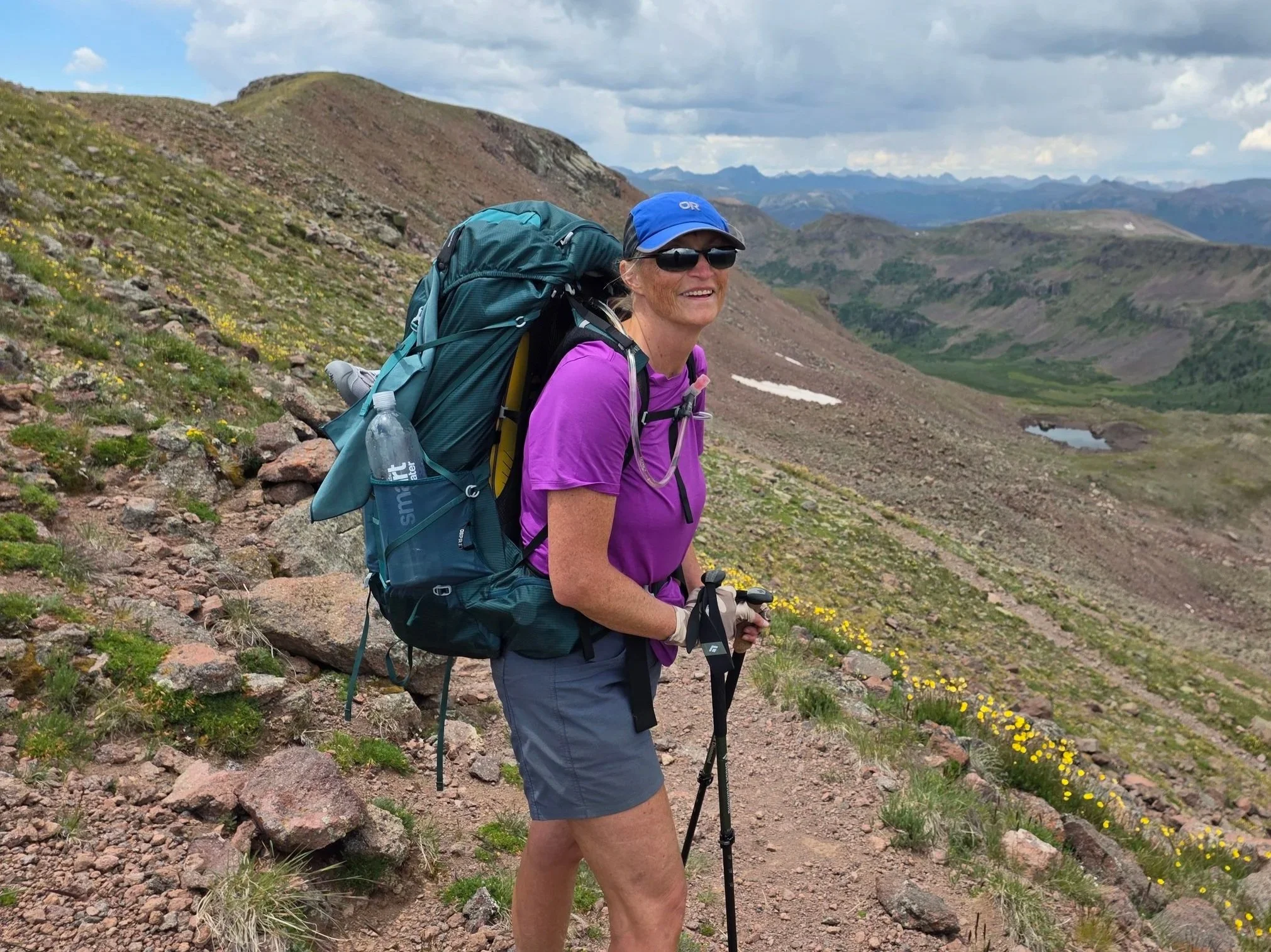 A woman hiking on a rocky trail in a mountainous landscape with green hills, patches of snow, and cloudy skies, smiling and wearing outdoor gear including a blue cap, sunglasses, a purple shirt, and carrying a large backpack.