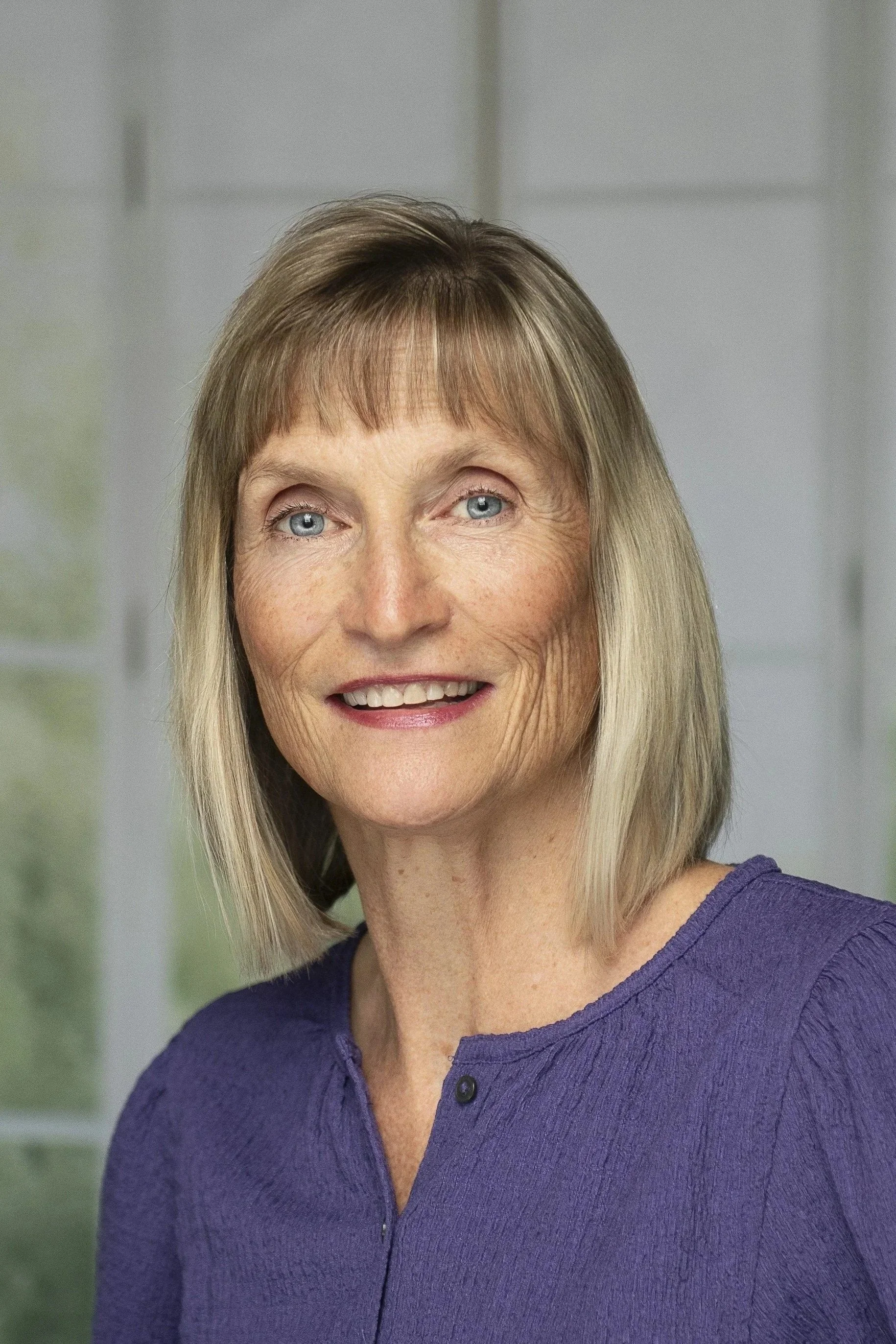 Senior woman with blonde hair, blue eyes, wearing a purple top, smiling, in a bright indoor setting.