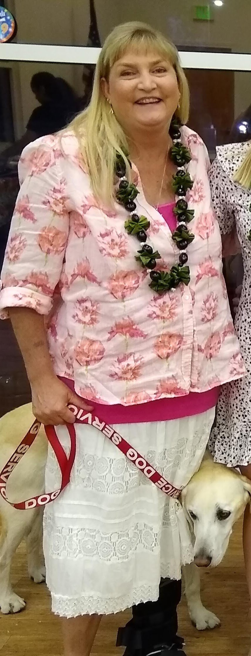 A smiling woman wearing a pink floral blouse and a lei, standing next to a white Labrador retriever with a red leash. The dog appears calm, and she is inside a building with some reflections and a partial view of another person in the background.