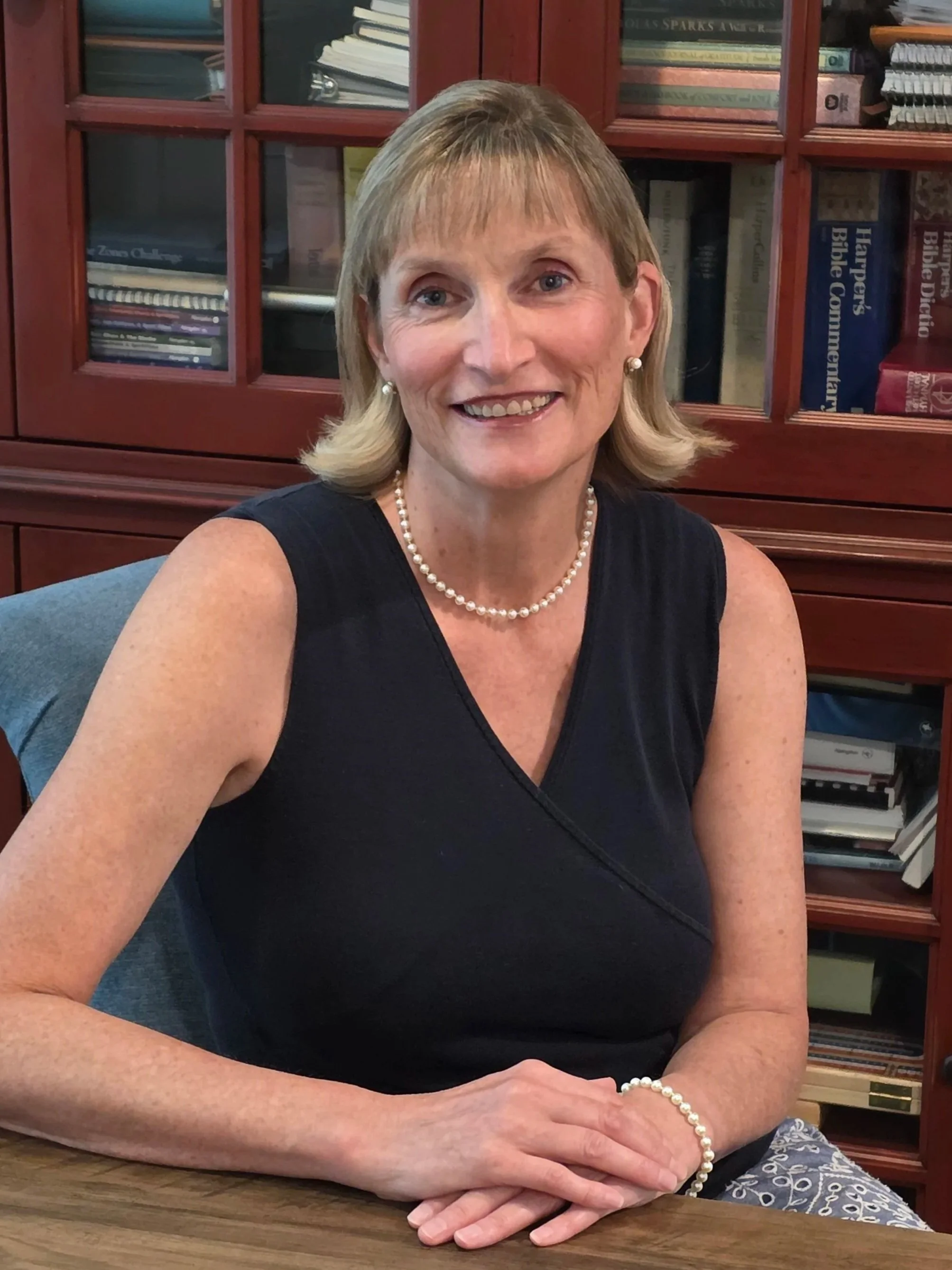 A middle-aged woman with blonde hair wearing a black sleeveless top, pearl earrings, and a pearl bracelet, sitting at a wooden table in front of a bookcase filled with books.