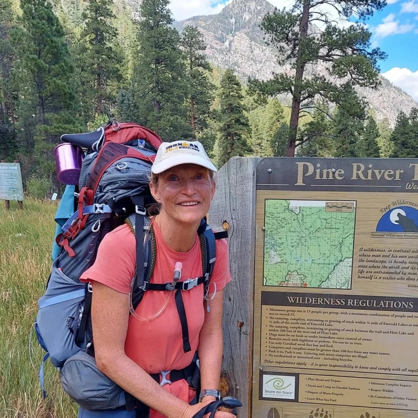 Smiling woman with a backpack standing next to a Pine River trail sign in a forested mountainous area.