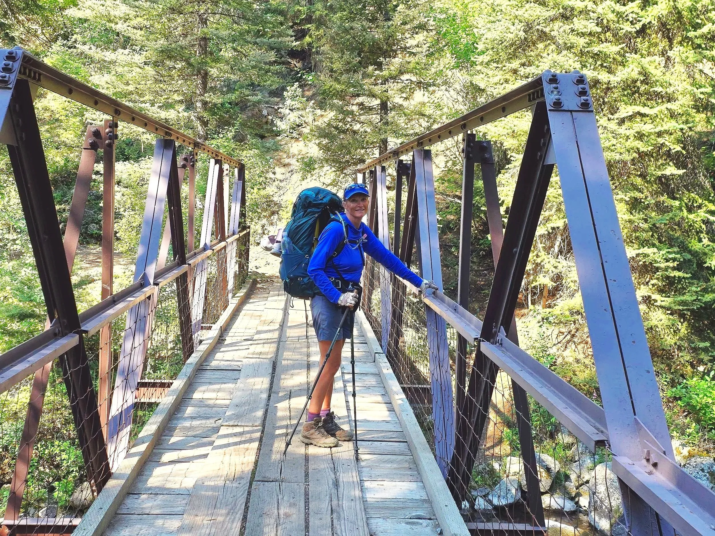 A woman with a blue jacket, grey shorts, hiking boots, and a large backpack standing on a wooden suspension bridge in a lush green forest, smiling at the camera.