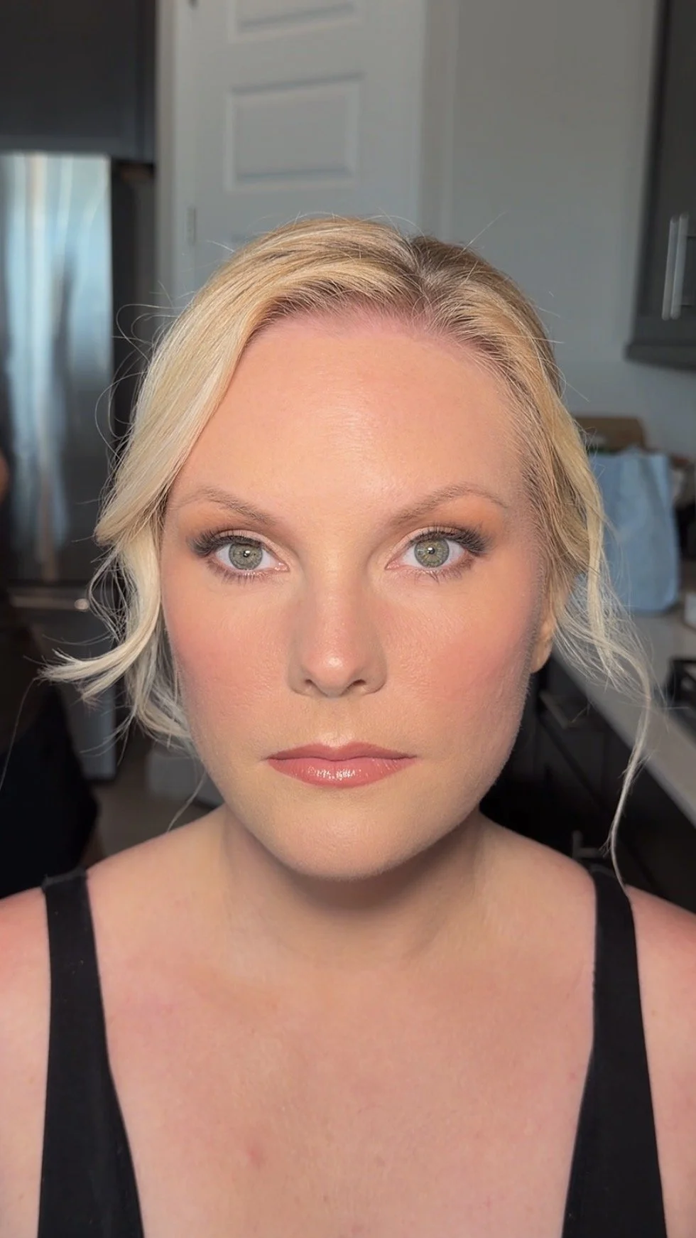 Close-up of a woman with short blonde hair and light makeup, wearing a black tank top, in a room with kitchen cabinets and a refrigerator in the background.