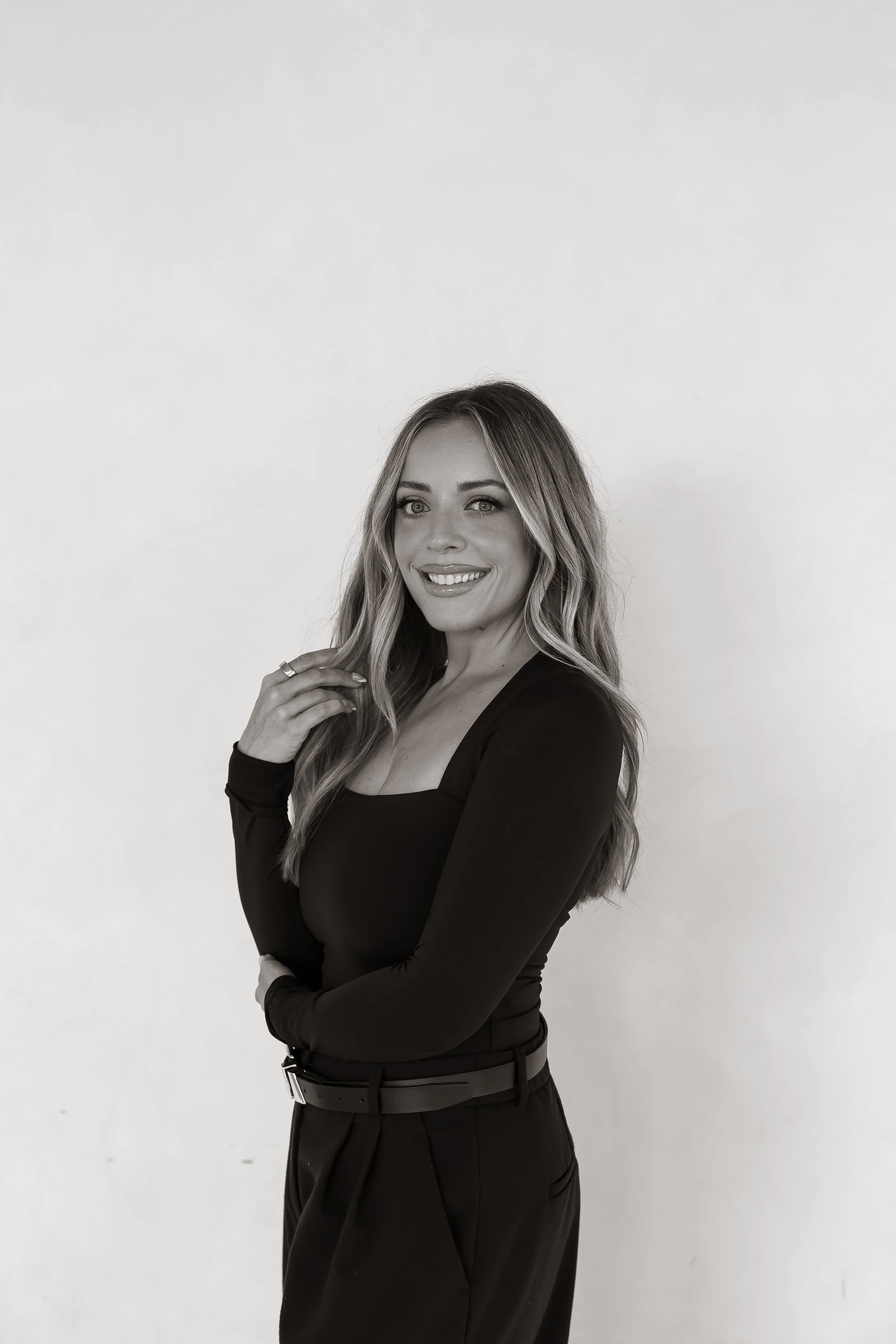 A smiling woman with long wavy hair wearing a long sleeve black top and black pants, standing against a plain white background.