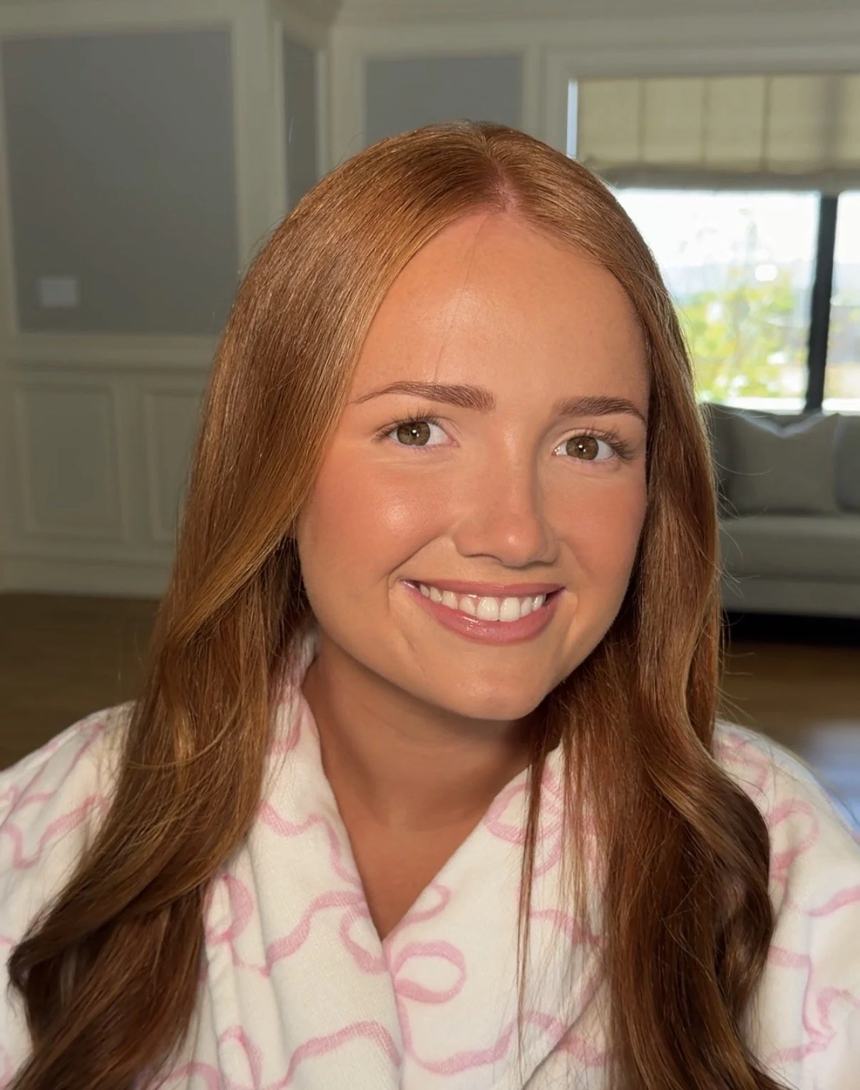 A young woman with long red hair smiling in a well-lit living room with a white sofa and large windows in the background.