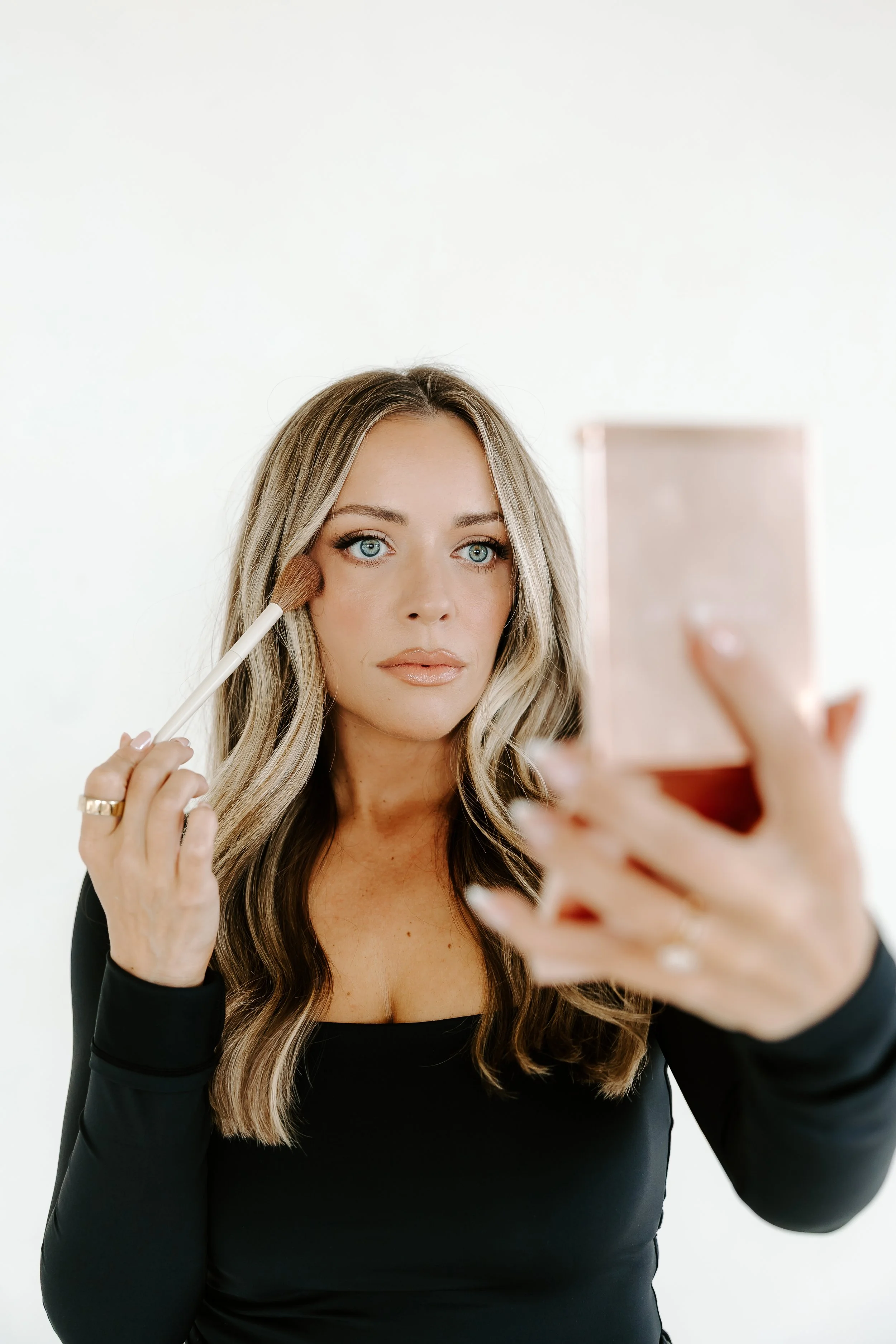 woman with long blonde hair applying makeup with brush while taking selfie in front of mirror