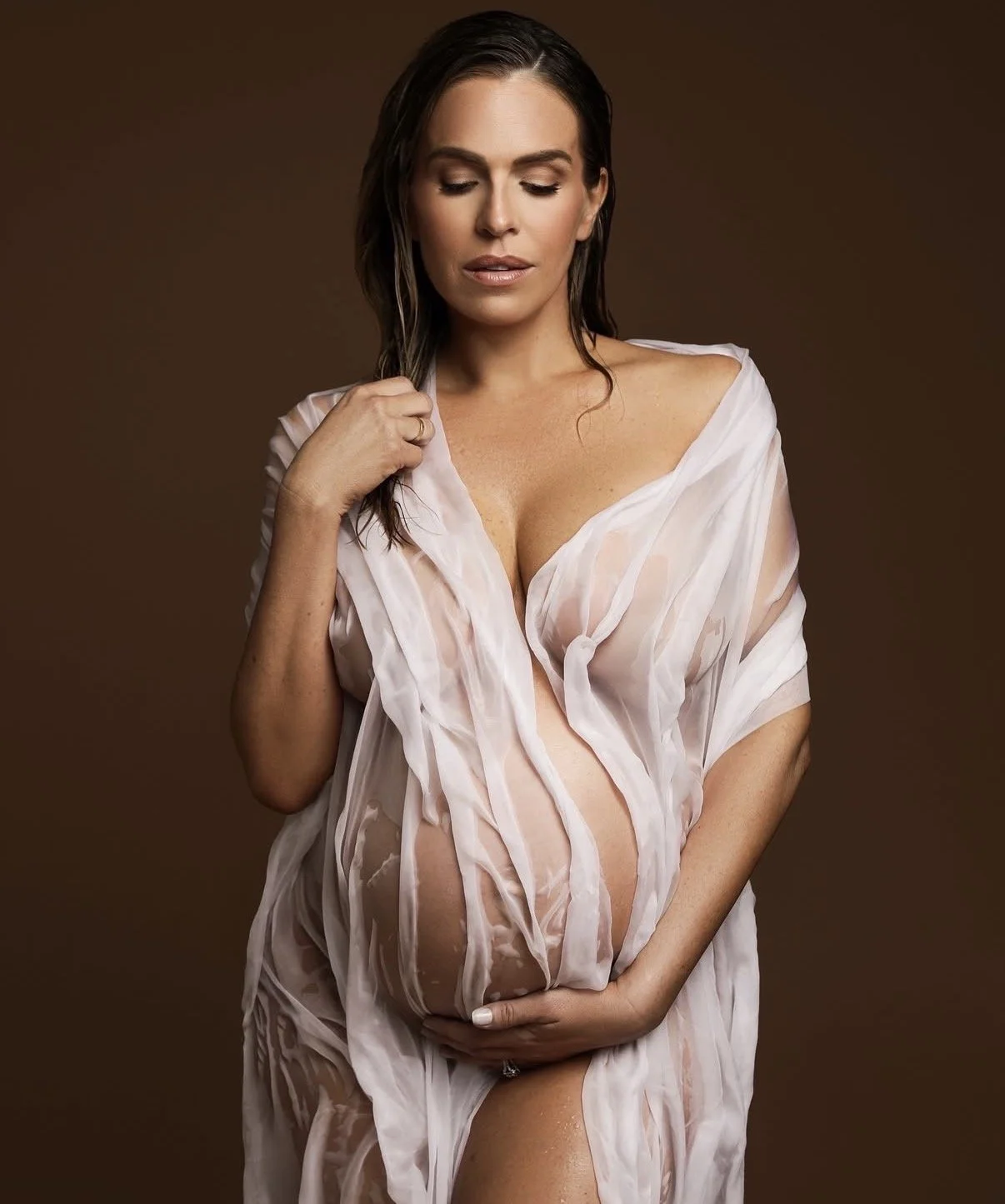 A woman with wet hair wearing a semi-transparent, white, wet dress against a brown background.
