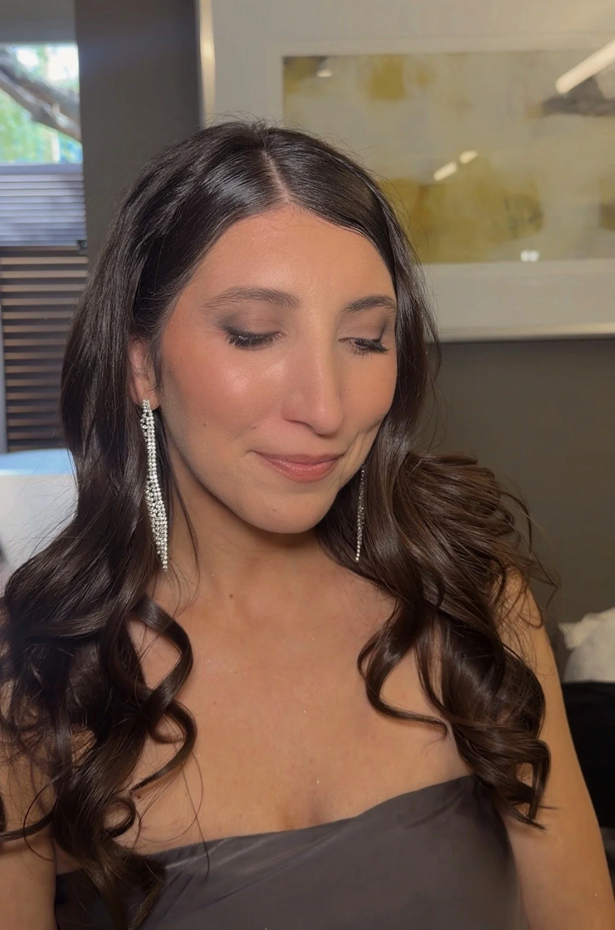 A woman with long, dark, curly hair wearing long, sparkly earrings and a strapless, dark gray dress, with her eyes closed and a slight smile.