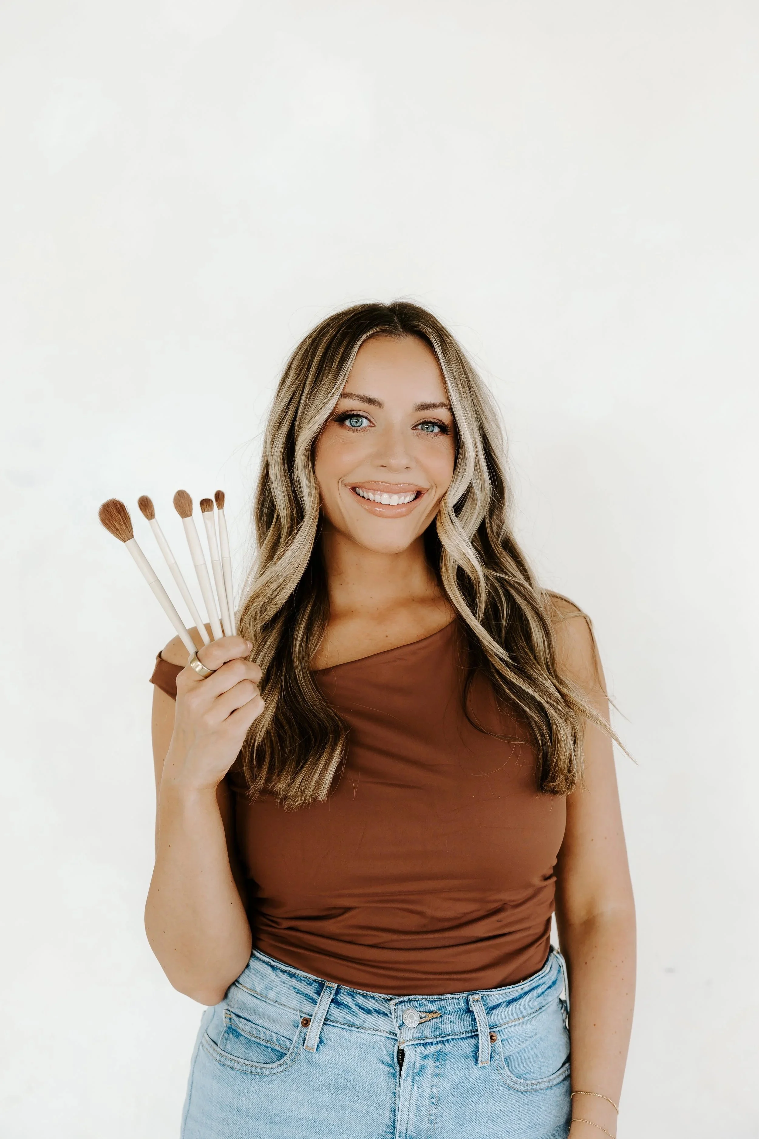A young woman with long, wavy blonde hair, smiling and holding a set of five makeup brushes against a white background. She is wearing a brown sleeveless top and light blue jeans.