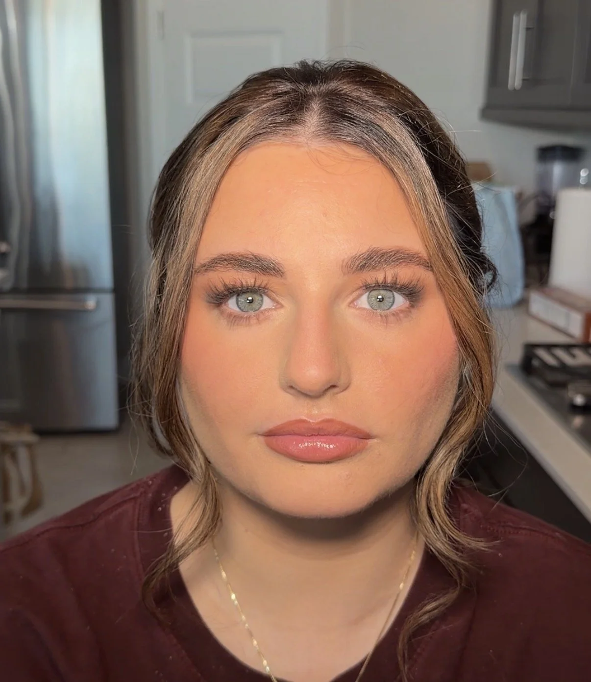 Close-up of a woman with light skin, shoulder-length wavy brown hair, bright blue eyes, and makeup, in a kitchen with appliances and cabinets in the background.