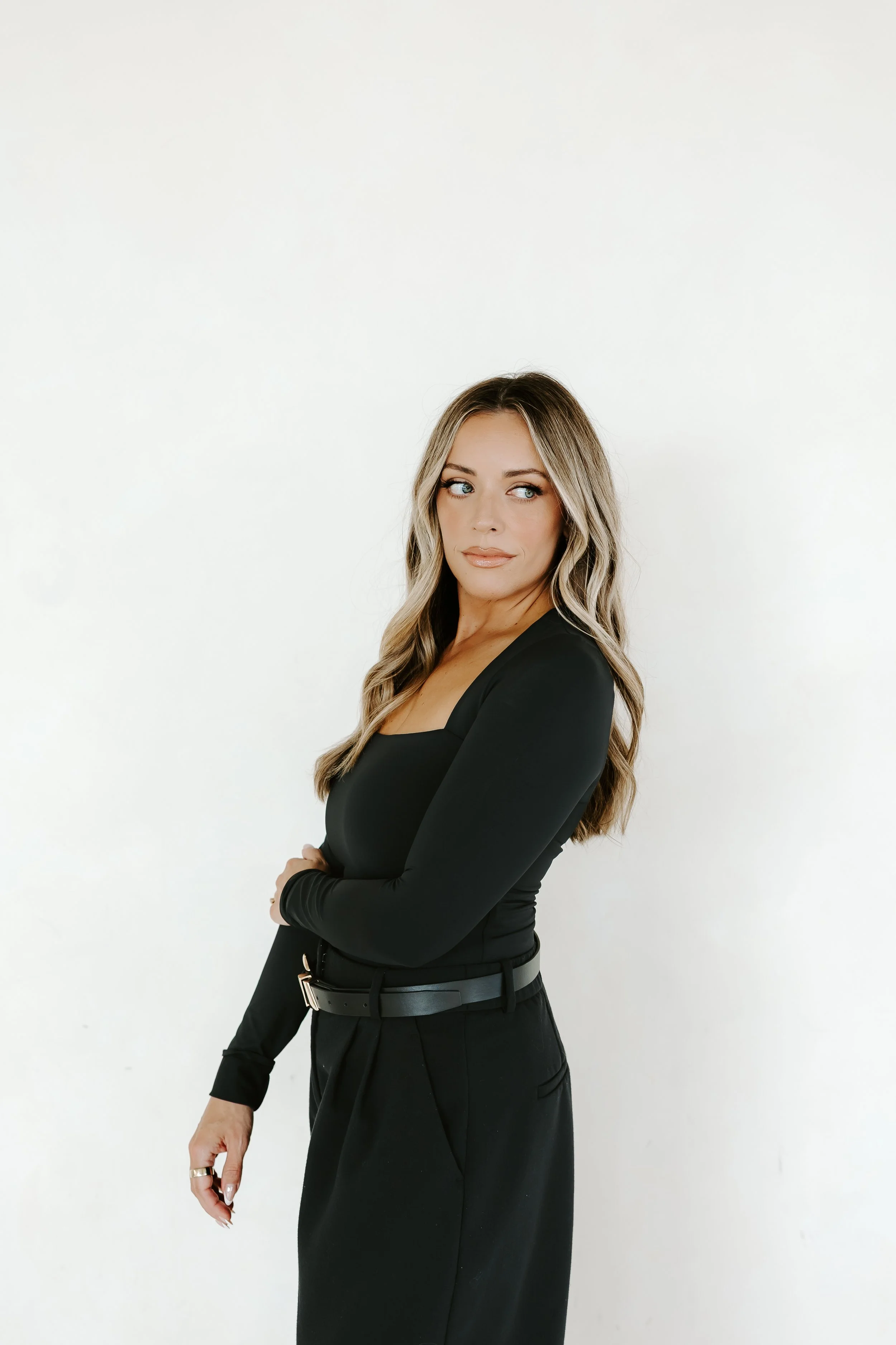 A woman with long wavy hair wearing a black long sleeve top and black pants standing against a plain white wall.