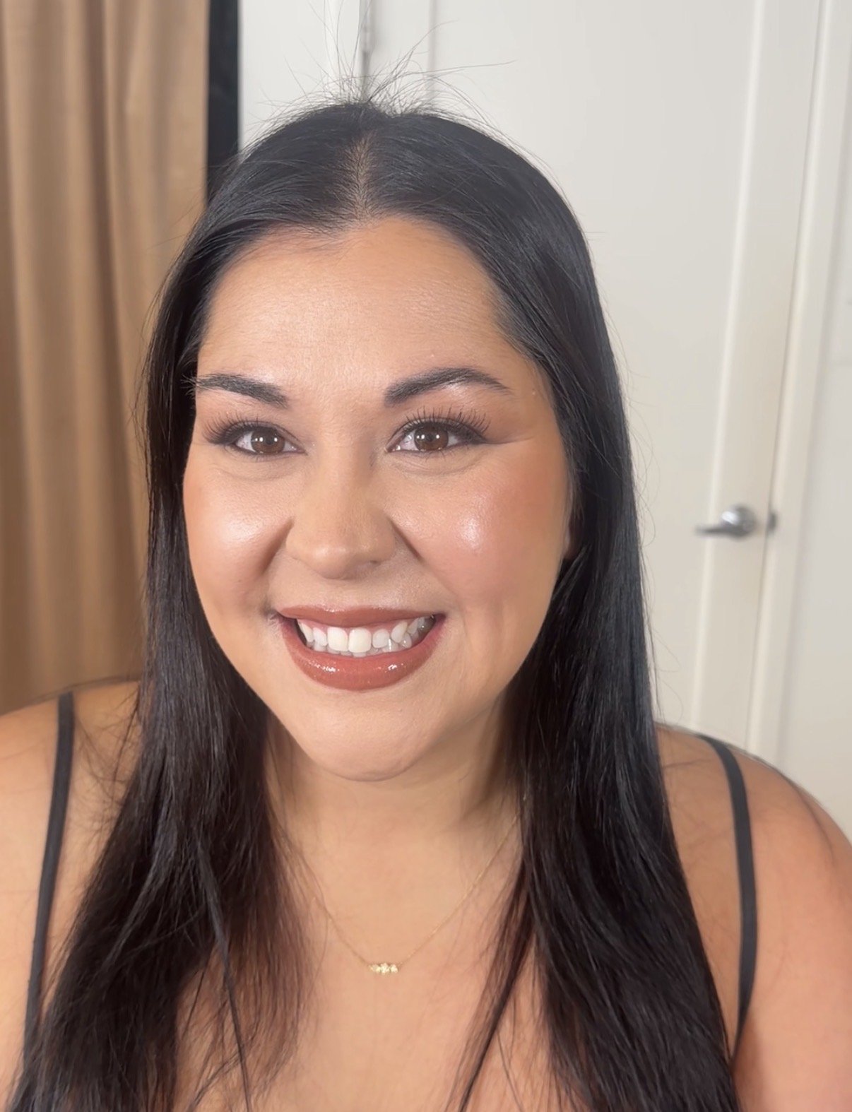 A smiling woman with long black hair, wearing makeup, a black tank top, and a gold necklace, standing indoors with a wall and door in the background.