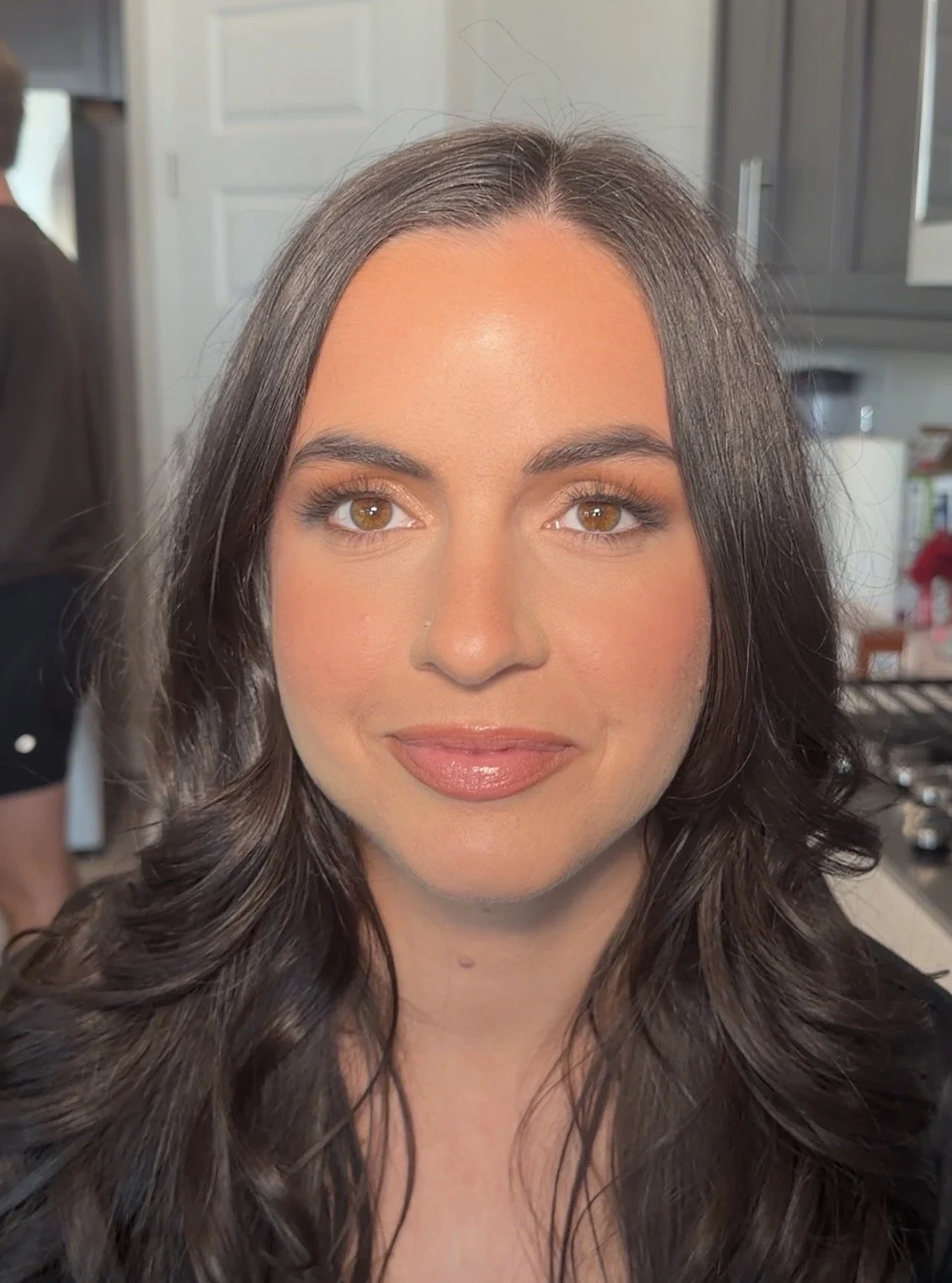 Close-up of a woman with long wavy brown hair, makeup, and brown eyes, smiling softly in a kitchen.