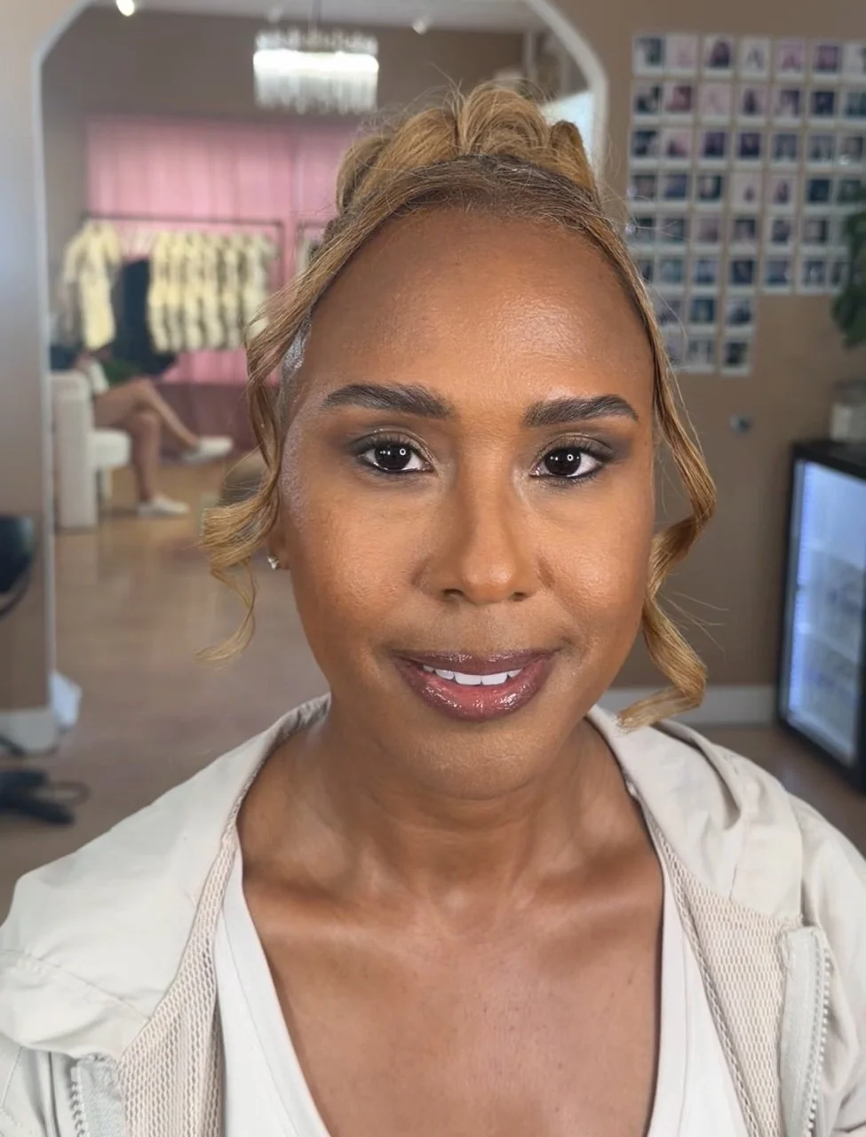 Close-up of a smiling woman with styled hair and makeup, indoors with photos on the wall and a person sitting in the background.